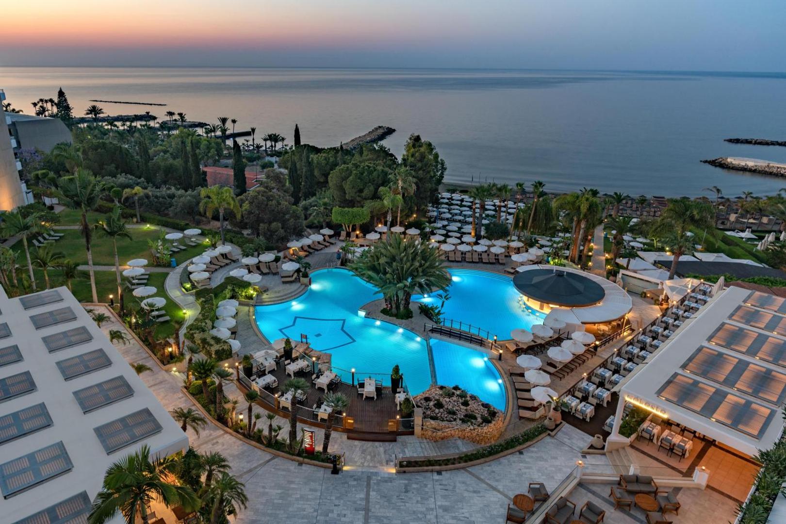 Mediterranean Beach Hotel teritorija saulrieta laikā — izgaismoti baseini, bārs un restorāna terase Limasolas piekrastē