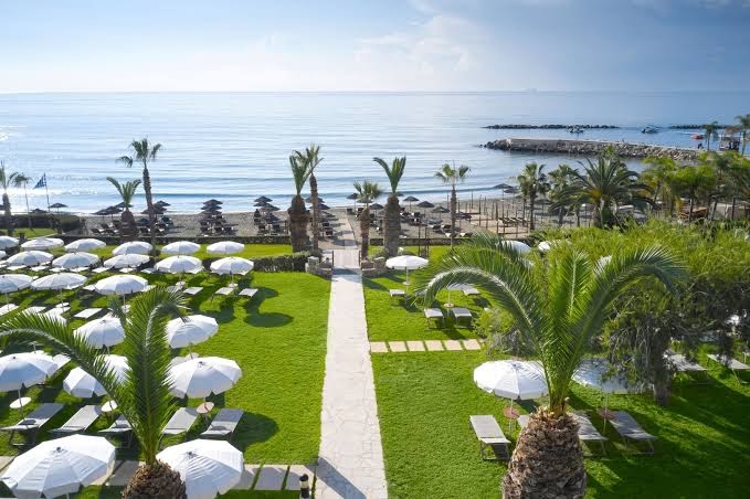 Mediterranean Beach Hotel zāliens ar saulessargiem un guļamkrēsliem pie pludmales — skats uz jūru, palmām un piekrastes zonu
