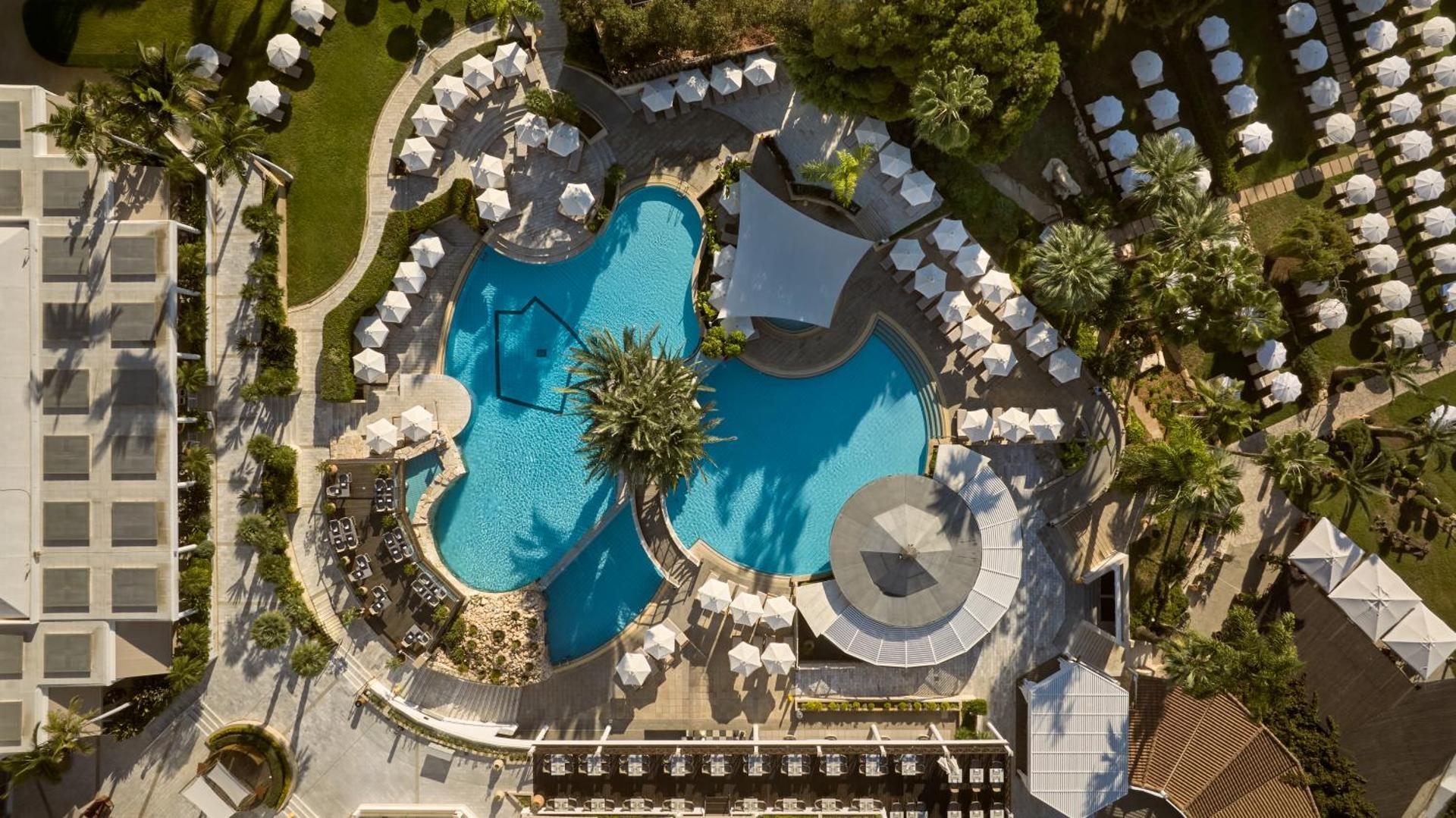 Mediterranean Beach Hotel baseinu zonas skats no putna lidojuma — zili baseini, balti saulessargi un palmas sakoptā dārzā