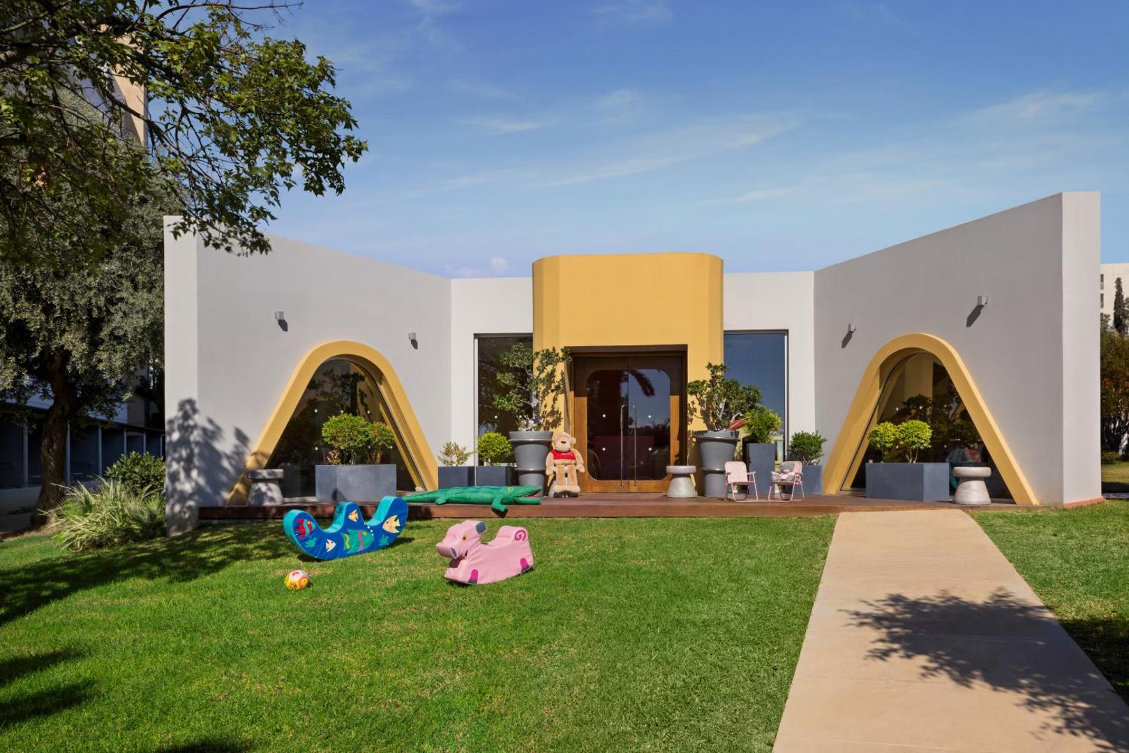Mediterranean Beach Hotel bērnu kluba ēka ar dzeltenām arkām un rotaļu laukumu zālienā — mūsdienīga arhitektūra un vieta āra aktivitātēm