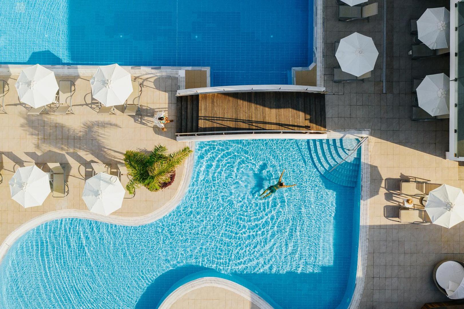 Sofianna Resort & Spa skats no augšas uz āra baseiniem Pafā, Kiprā — koka tilts, balti saulessargi un atpūtas zona