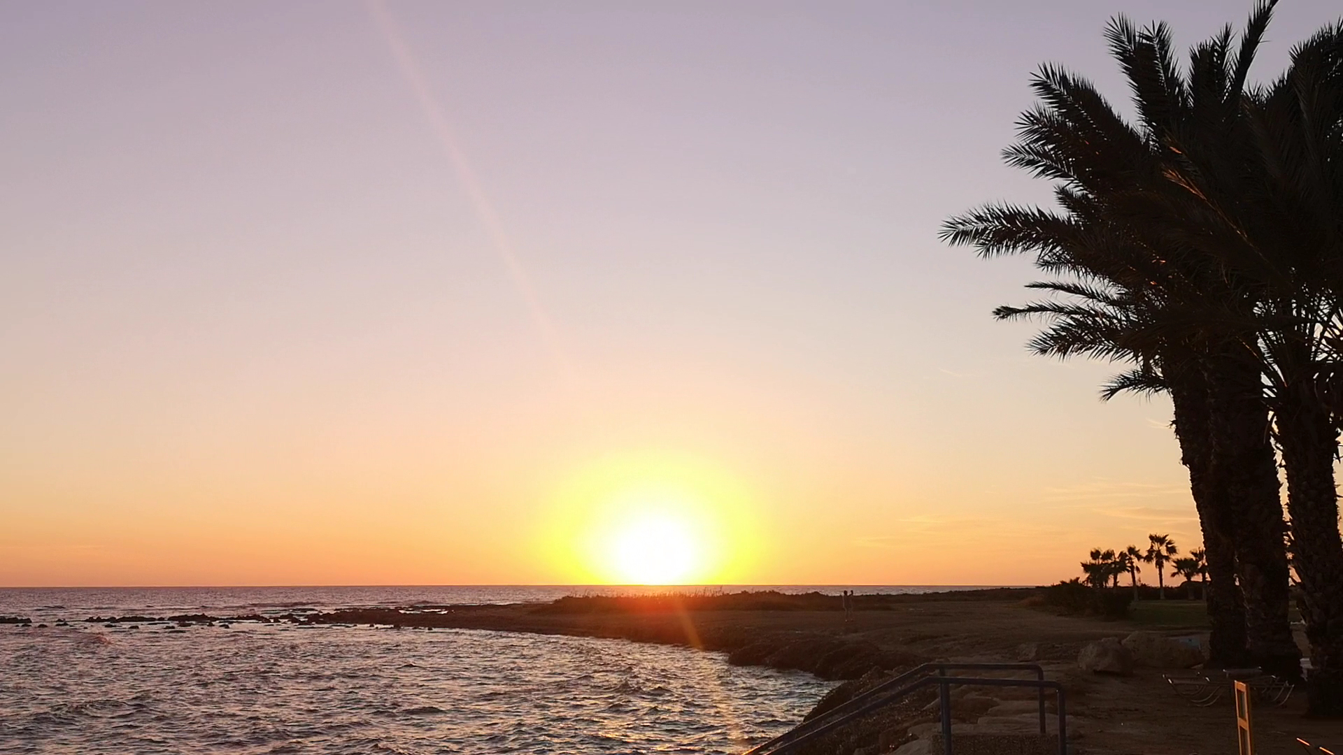 Saulriets pie Louis Phaethon Beach viesnīcas Pafā — skats uz Vidusjūras piekrasti un palmu siluetiem vakara saules gaismā