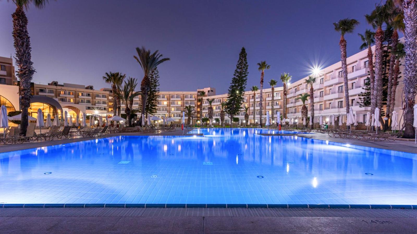 Viesnīcas Louis Phaethon Beach korpusi un izgaismots āra baseins vakarā — plaša atpūtas zona ar palmām un zviļņiem