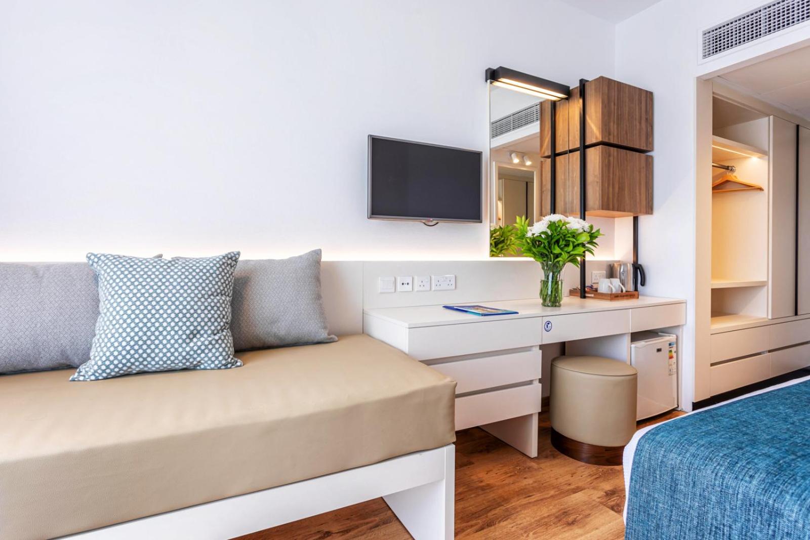 Louis Phaethon Beach moderns viesnīcas numurs ar gaišu interjeru, dīvānu, rakstāmgaldu un TV — mājīga atpūtas zona Pafā