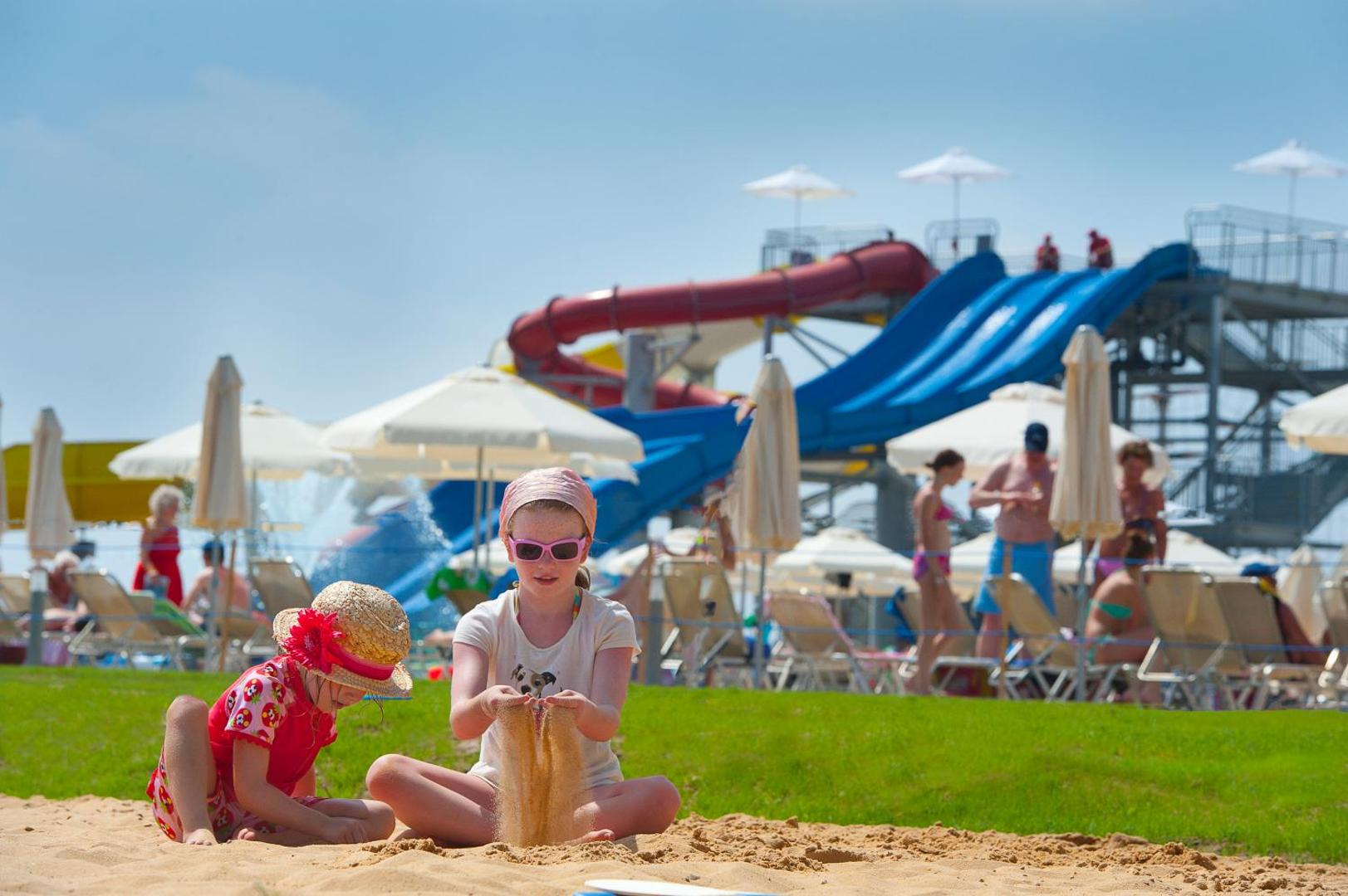 Bērni spēlējas smiltīs Louis Phaethon Beach akvaparka fonā Pafā — redzami lieli ūdens slidkalniņi un sauļošanās zona ar saulessargiem