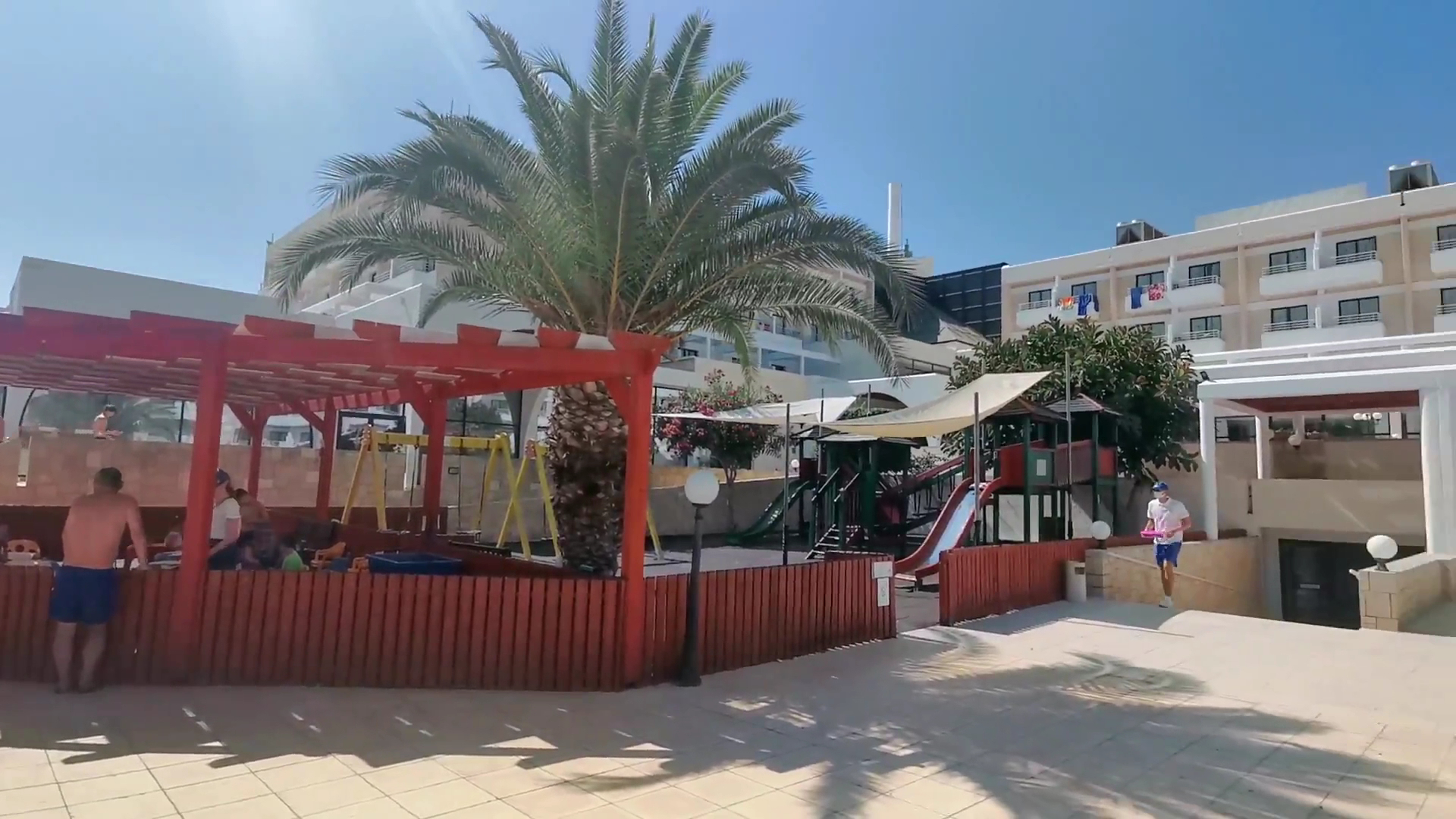 Louis Phaethon Beach bērnu rotaļu laukums ar slidkalniņiem un šūpolēm zem palmas — blakus viesnīcas korpuss un atpūtas nojume