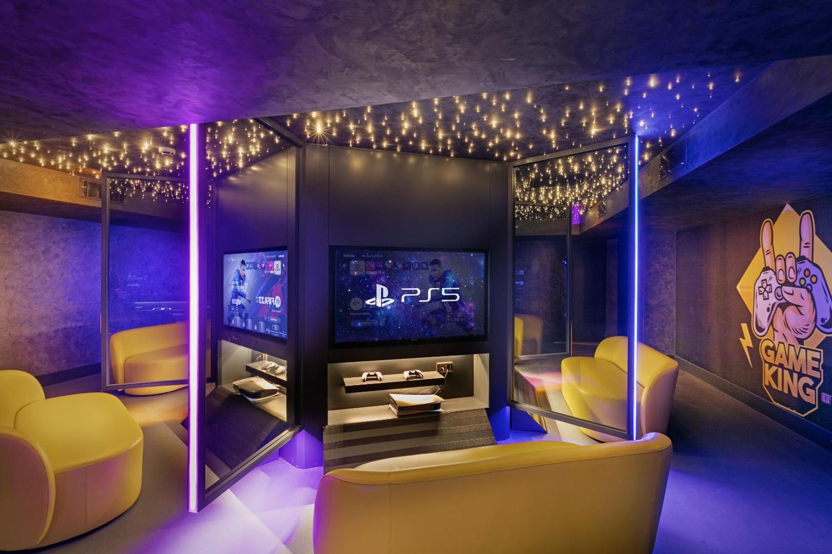Titanic Deluxe Lara spēļu telpa pusaudžiem ar PlayStation 5 konsolēm, ērtiem dzelteniem dīvāniem un modernu neona apgaismojumu