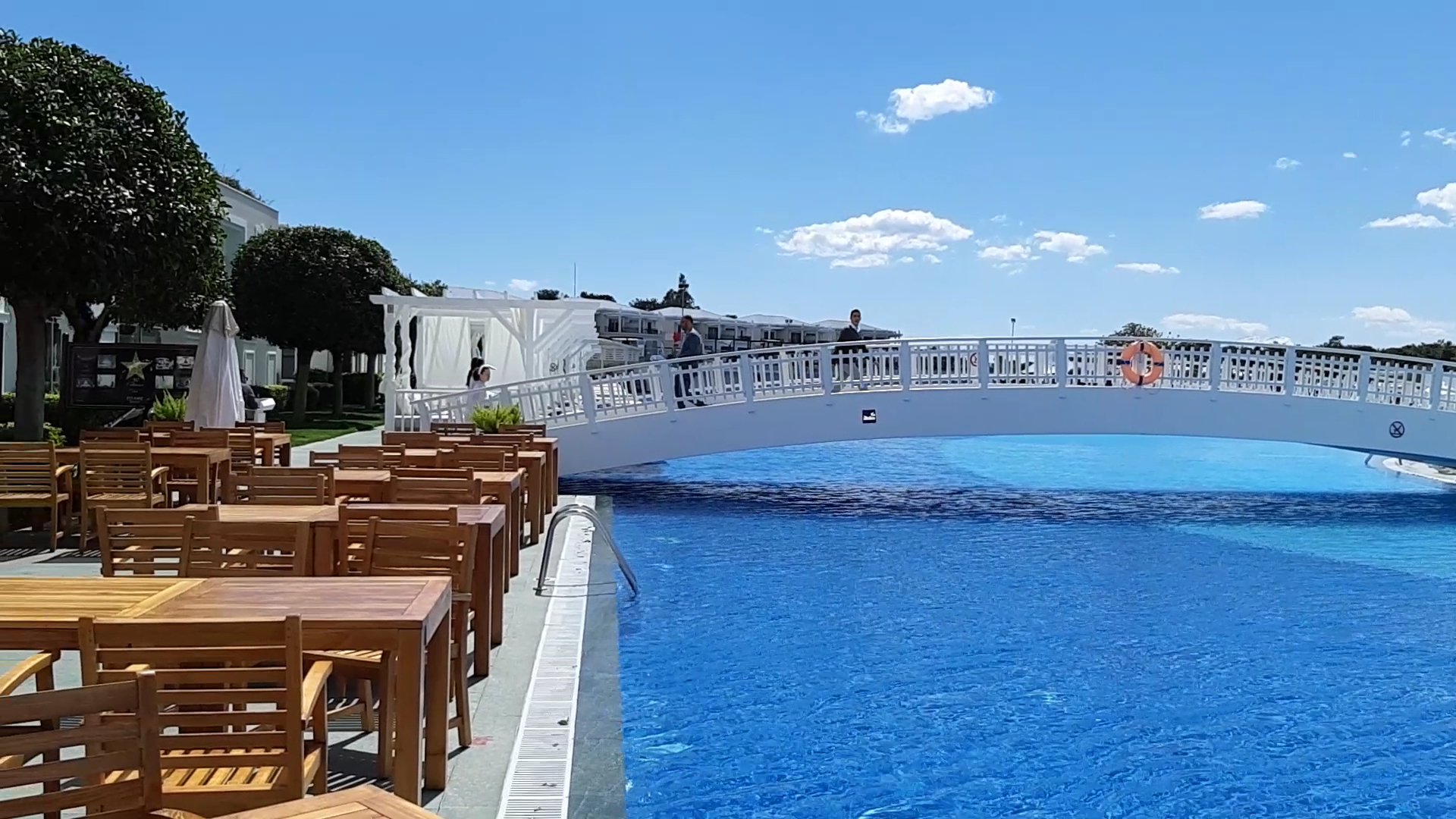 Titanic Deluxe Golf Belek āra terase ar koka galdiem pie baseina un balts tilts pāri ūdenim saulainā dienā Belekā