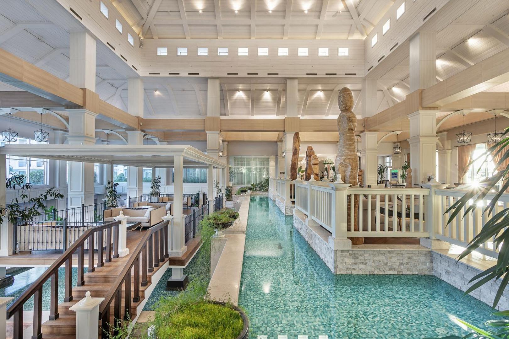 Titanic Deluxe Golf Belek plašais lobby interjers ar dekoratīvu ūdens kanālu, koka tiltiņiem un augstiem griestiem