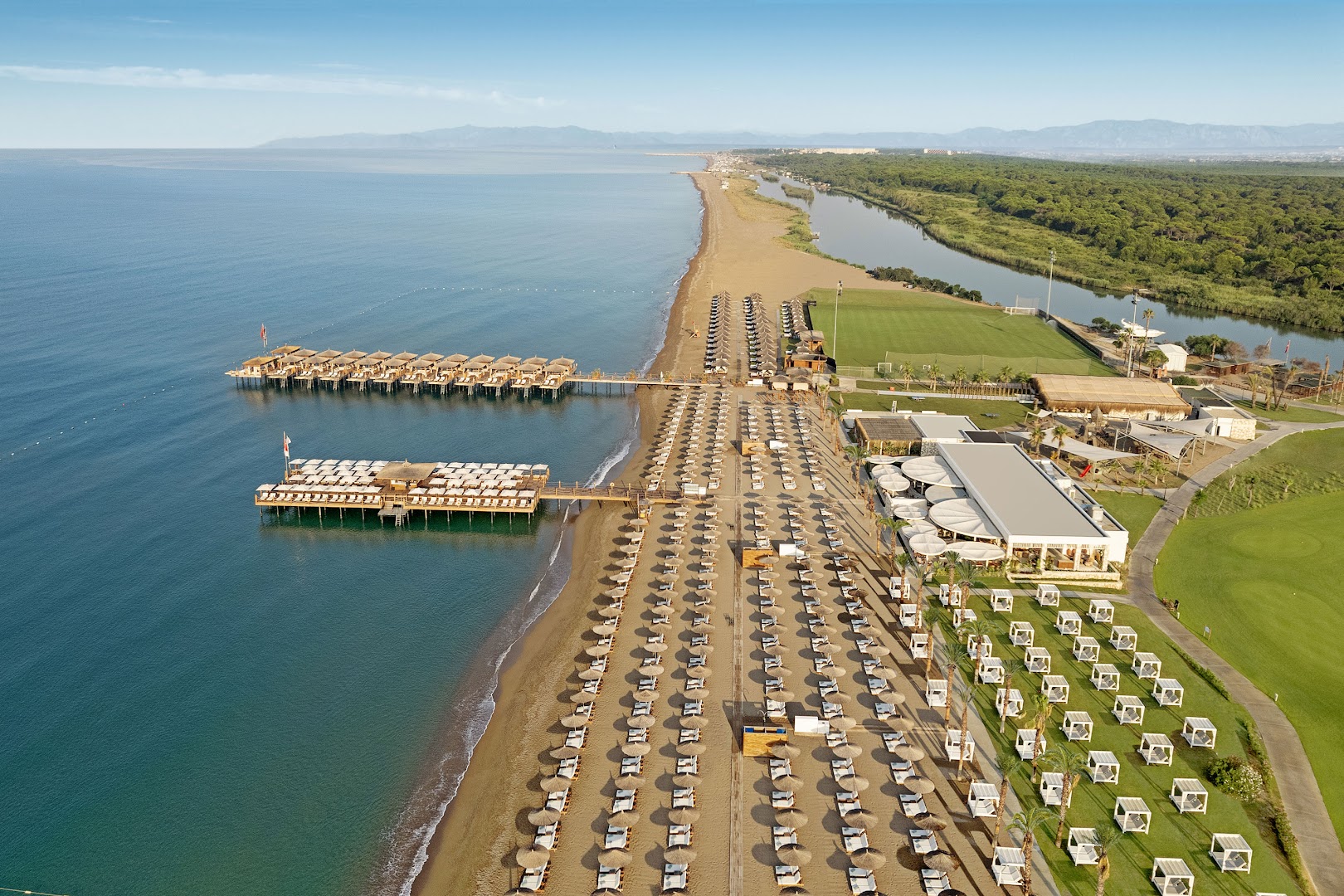 Aerofoto skats uz Titanic Deluxe Golf Belek pludmales zonu ar saulessargiem, gultām un divām koka laipām jūrā