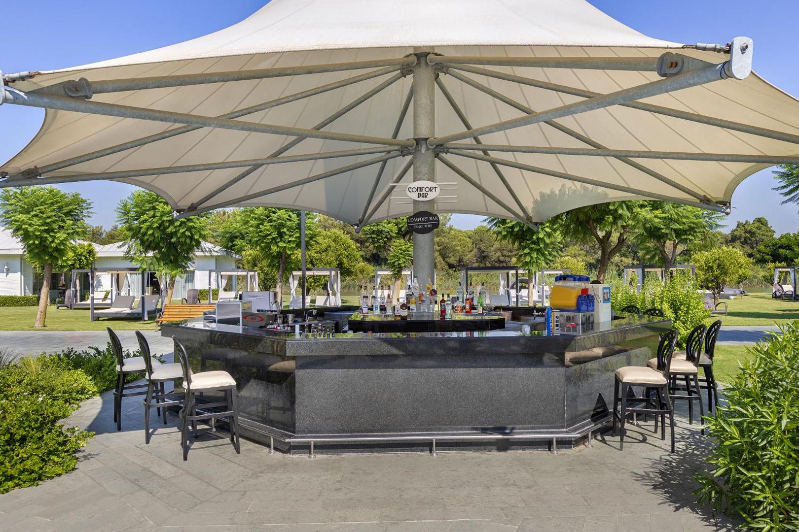 Titanic Deluxe Golf Belek Comfort Bar āra bārs dārza zonā — bāra lete ar dzērieniem zem liela saulessarga un atpūtas krēsliem