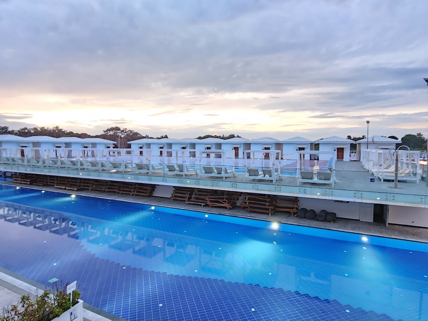 Titanic Deluxe Golf Belek peldbaseins saulrieta laikā ar apgaismojumu un atpūtas zonām — skats uz baseina terasi un bungalo