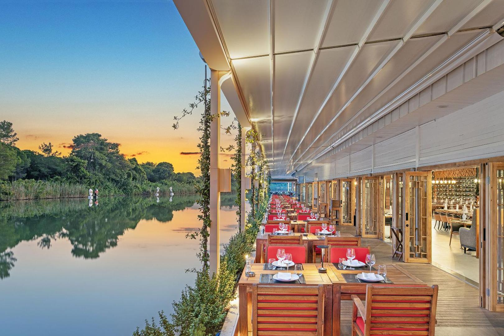 Titanic Deluxe Golf Belek āra restorāna terase pie upes ar klātiem galdiem un sarkanu akcentu krēsliem vakara gaismā