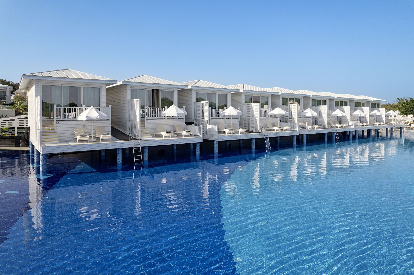 Gaiši koka bungalo uz pāļiem virs zila baseina ūdens viesnīcā Titanic Deluxe Golf Belek — privātas terases ar saulessargiem