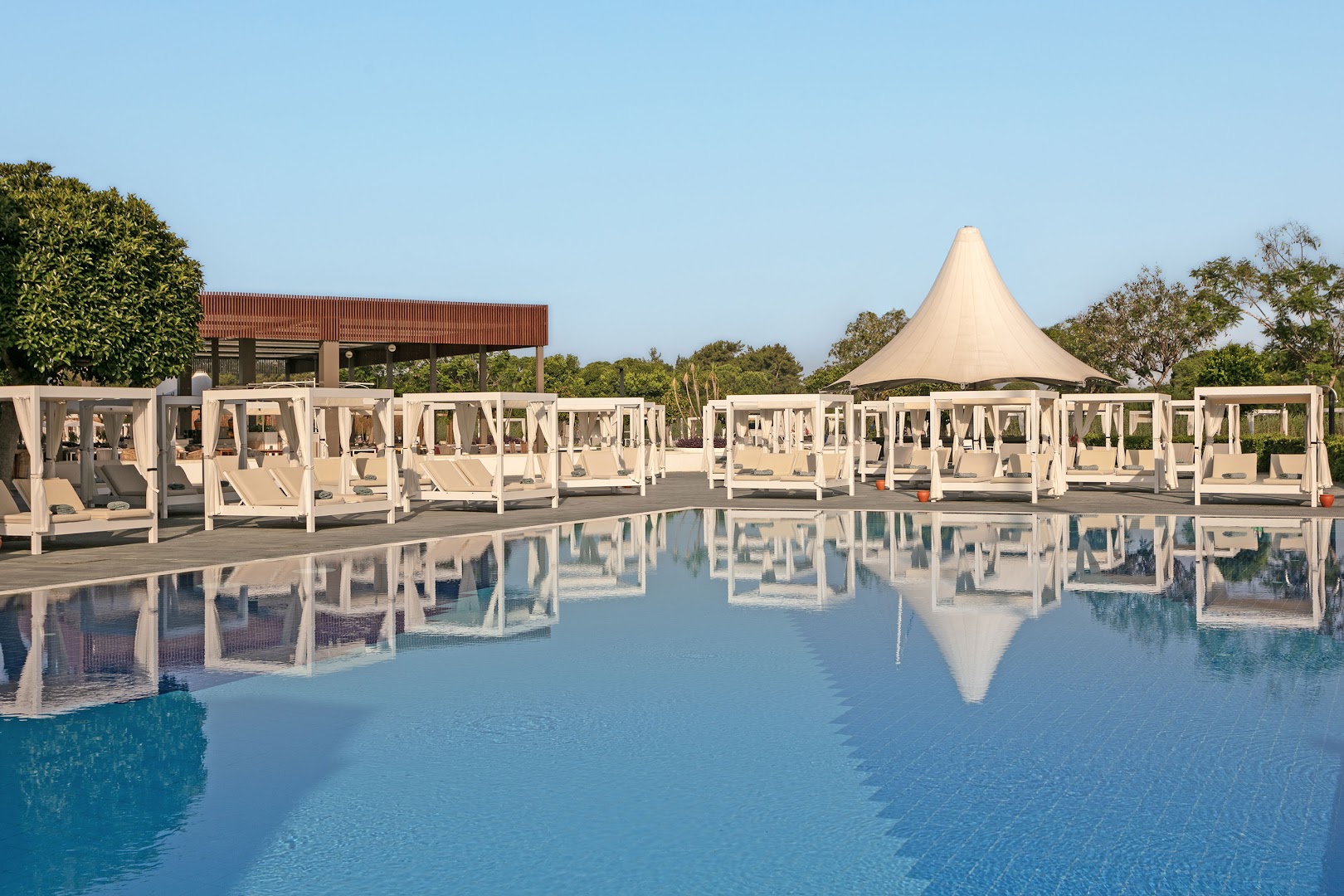 Titanic Deluxe Golf Belek atpūtas zona ar baltām nojumēm pie baseina — grezni sauļošanās krēsli un skats uz baseina spoguļvirsmu