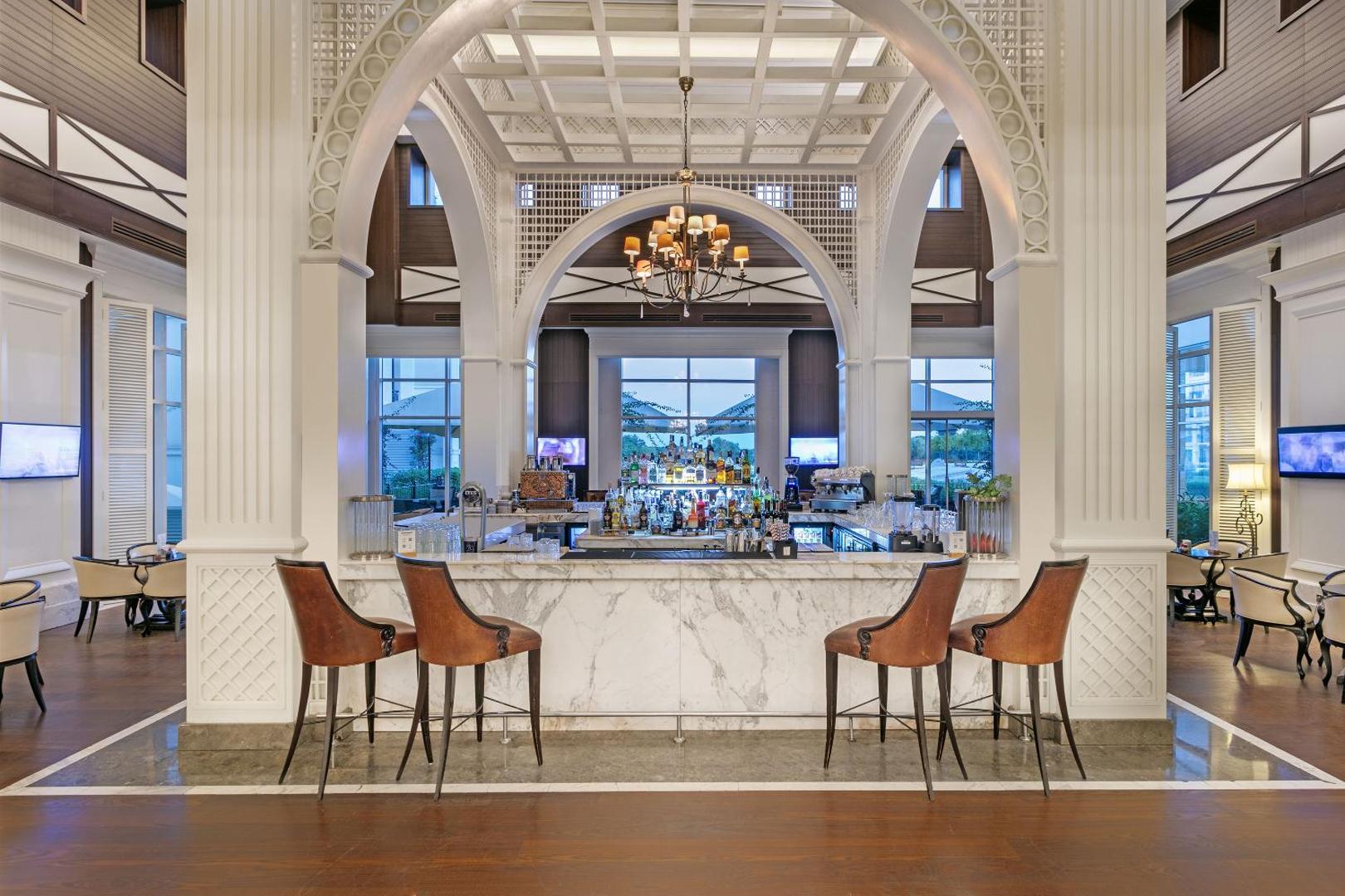 Titanic Deluxe Golf Belek viesnīcas lobby bārs ar marmora leti, greznu lustru un klasiska stila ādas krēsliem elegantā interjerā