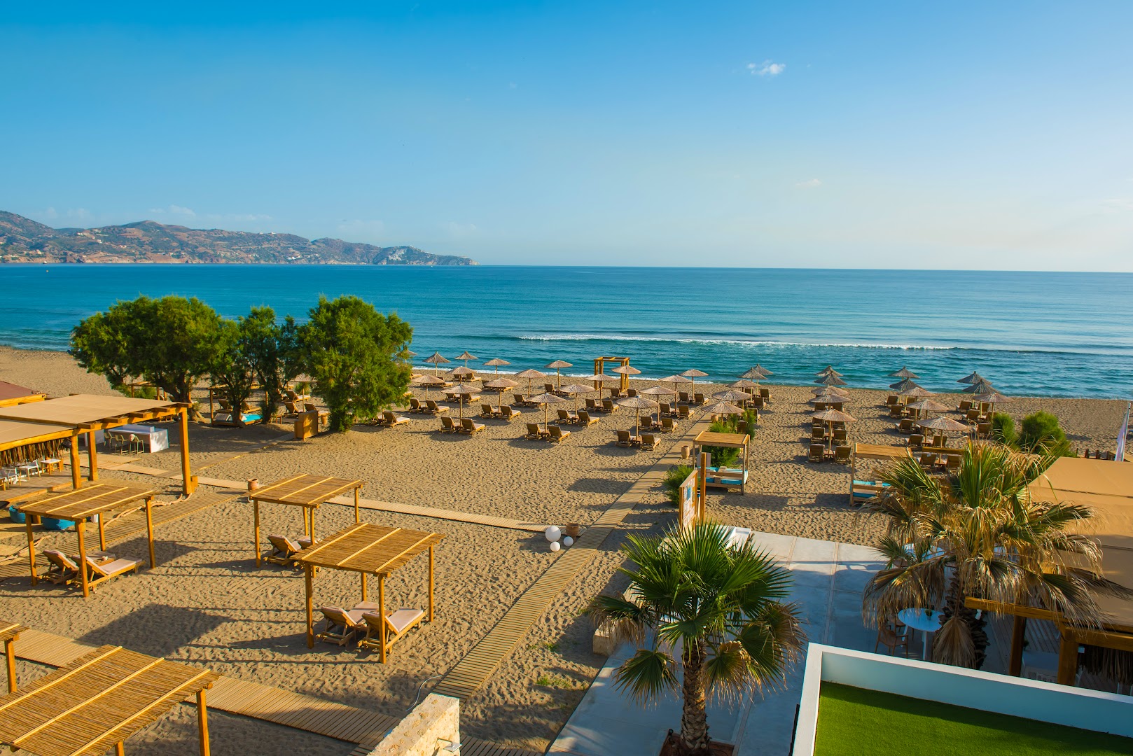 Plašs skats uz Paralos Lifestyle Beach pludmali un Vidusjūru — koka laipas, saulessargi un atpūtas zonas smilšainajā piekrastē