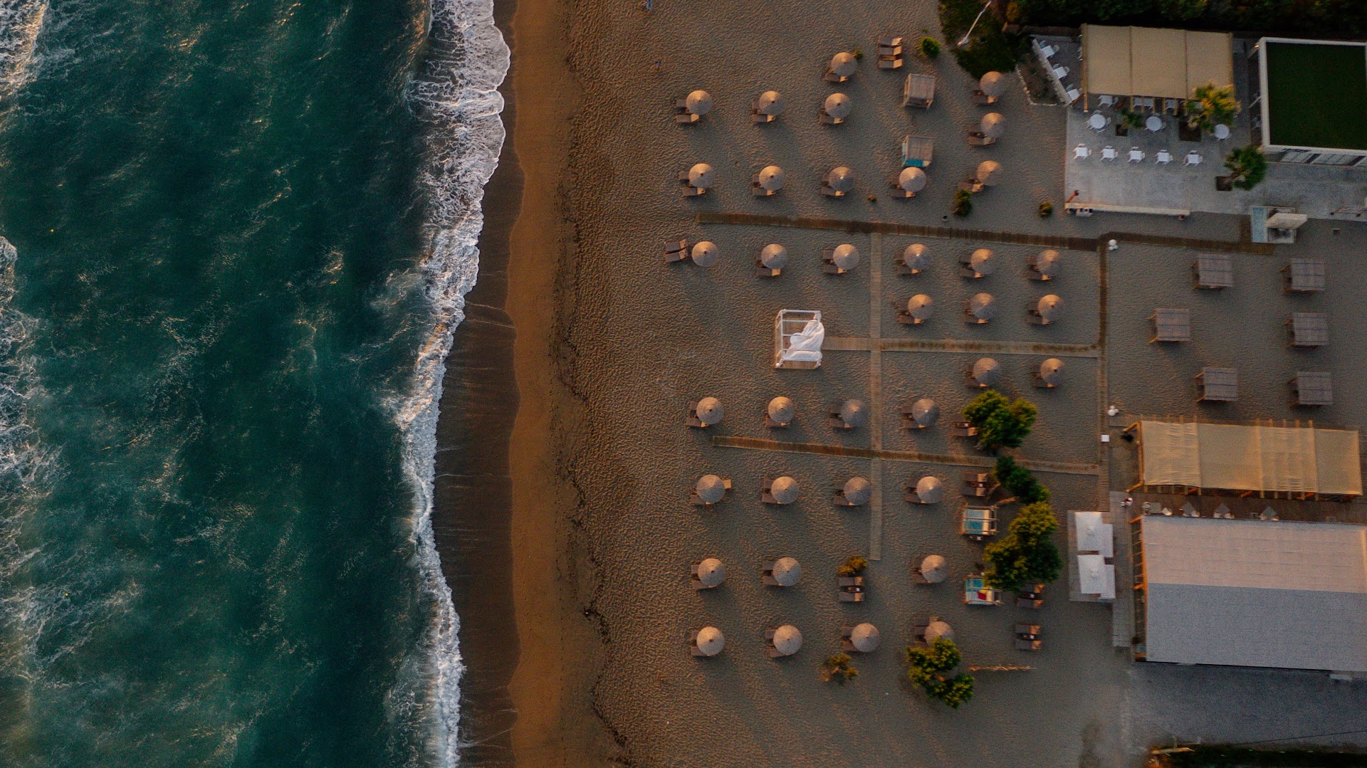 Aerofoto skats uz Paralos Lifestyle Beach privāto pludmali saulrietā — sakoptas atpūtas zonas ar saulessargiem un gultām pie jūras