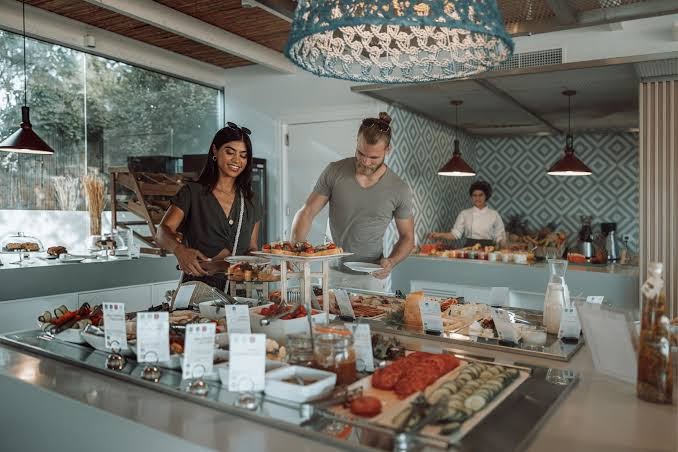 Viesi izvēlas ēdienus Paralos Lifestyle Beach viesnīcas bufetes restorānā — plaša uzkodu un svaigu produktu izvēle modernā interjerā