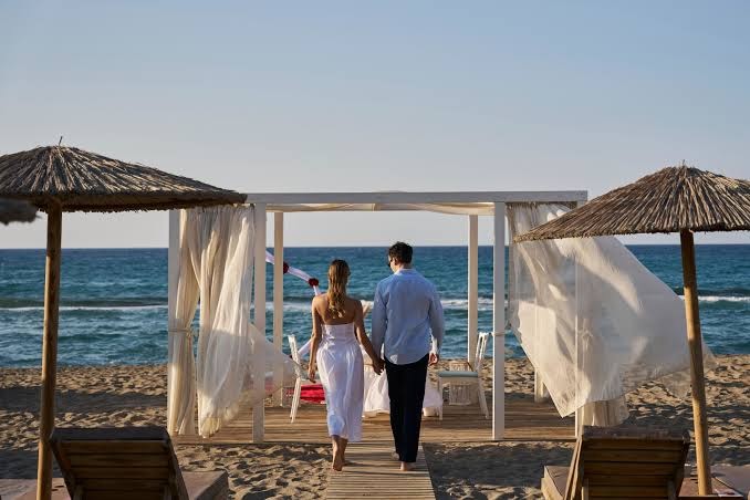 Pāris baltā apģērbā pastaigājas Paralos Lifestyle Beach pludmales zonā pie dekoratīvas nojumes — romantiska gaisotne pie jūras Krētā