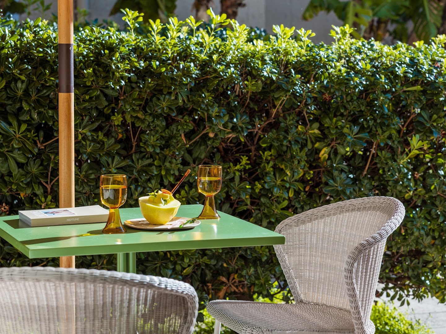 Grecotel Casa Adele āra terase ar zaļu galdiņu, pītu krēslu un svaigiem augļiem uz dārza fona saulainā dienā