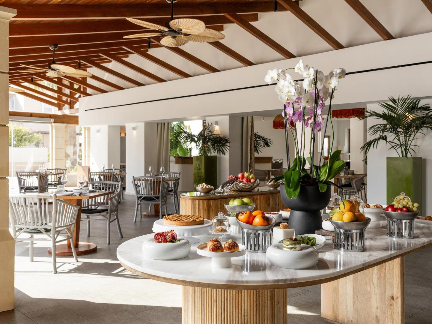 Grecotel Casa Adele restorāna brokastu bufete ar svaigiem augļiem un konditorejas izstrādājumiem uz marmora galda gaišā, modernā telpā
