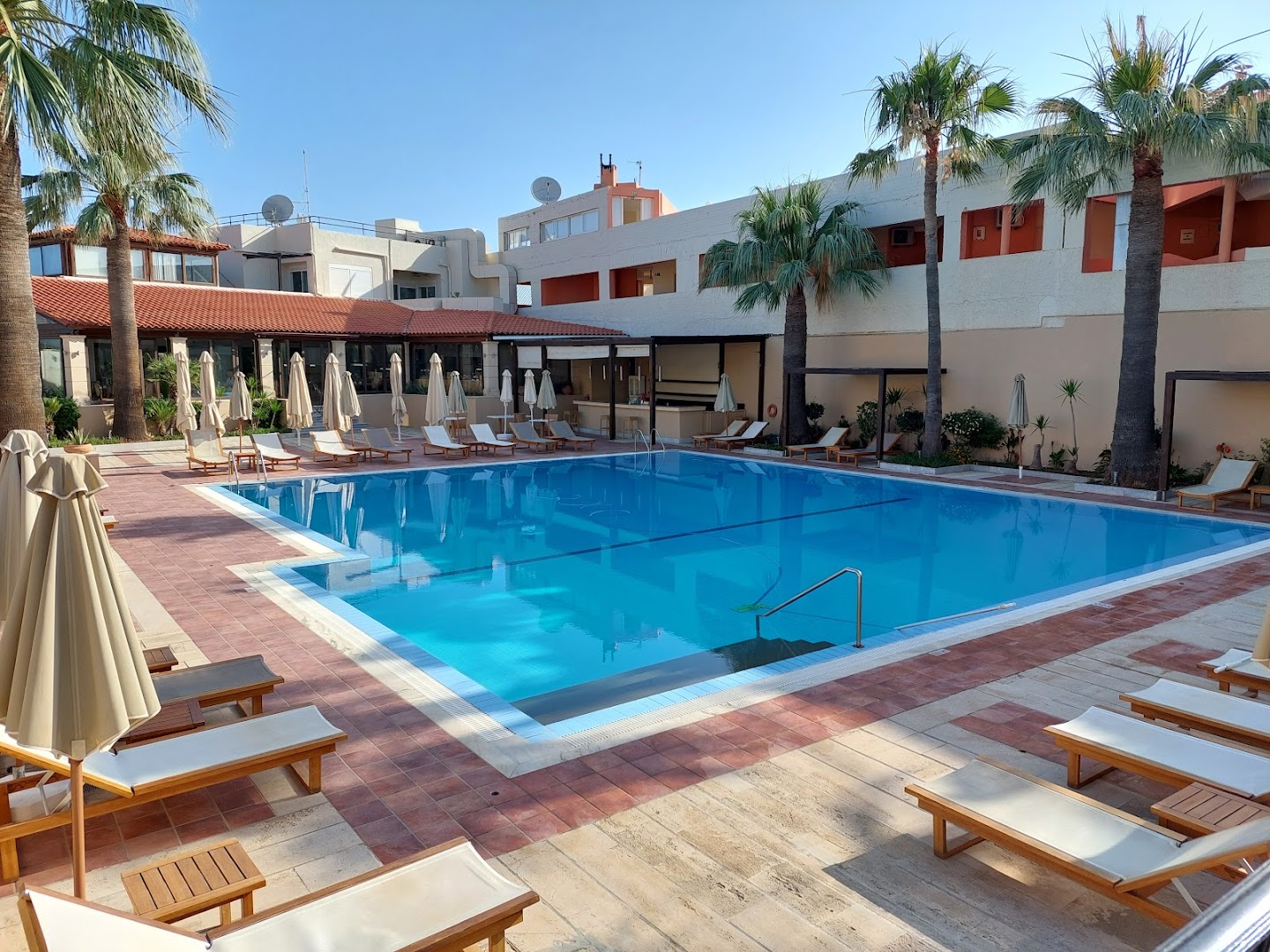 Grecotel Casa Adele viesnīcas iekšpagalms ar taisnstūra formas baseinu, koka sauļošanās krēsliem un bāru fonā
