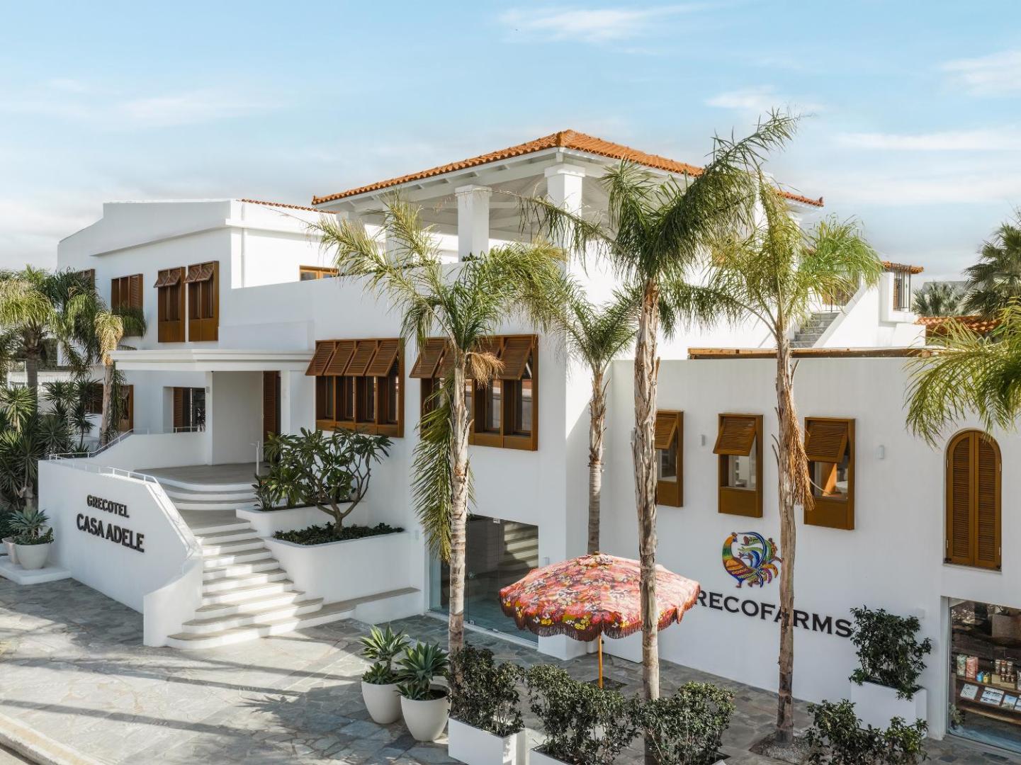 Grecotel Casa Adele viesnīcas galvenā ēka ar baltu fasādi, koka slēģiem un palmām Krētā, Grieķijā saulainā dienā