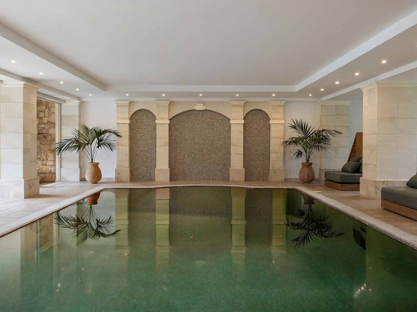Grecotel Casa Adele spa zonas iekštelpu baseins ar tirkīza ūdeni, akmens kolonnām un atpūtas krēsliem relaksējošā gaisotnē