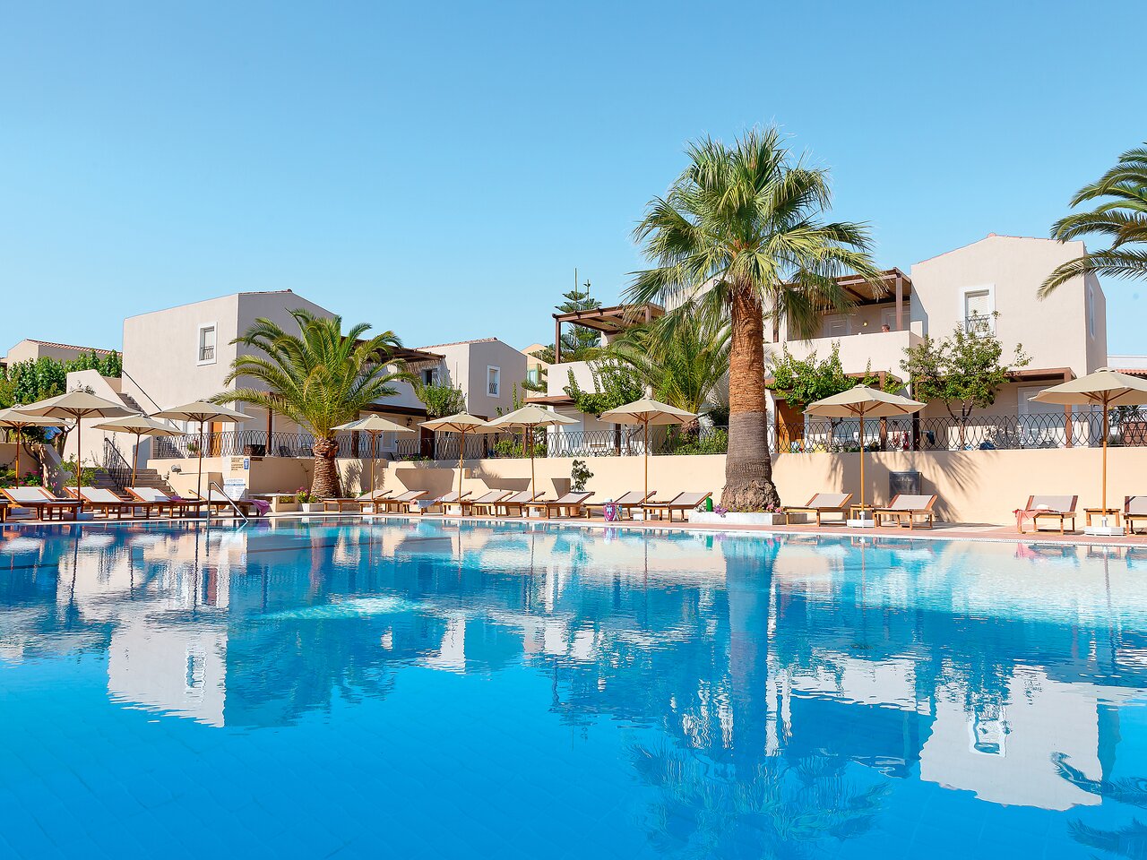 Grecotel Casa Adele āra baseins ar dzidru ūdeni, saulessargiem un palmām Krētā — skats uz viesnīcas korpusiem saulainā dienā