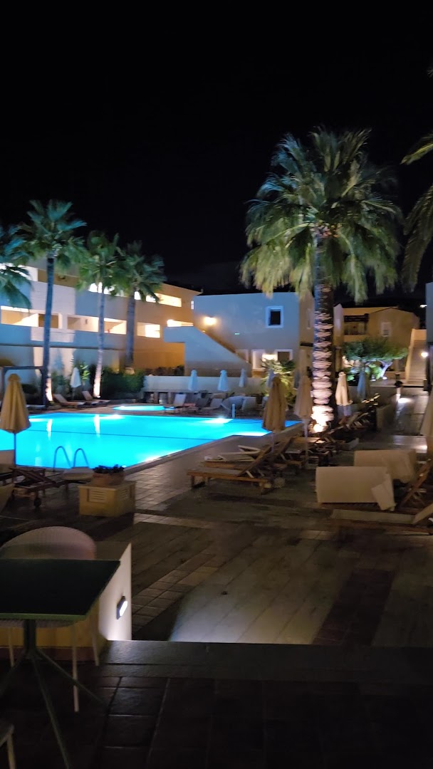Grecotel Casa Adele nakts skats uz izgaismotu baseinu un palmām — viesnīcas numuru korpusi fonā zem tumšām debesīm