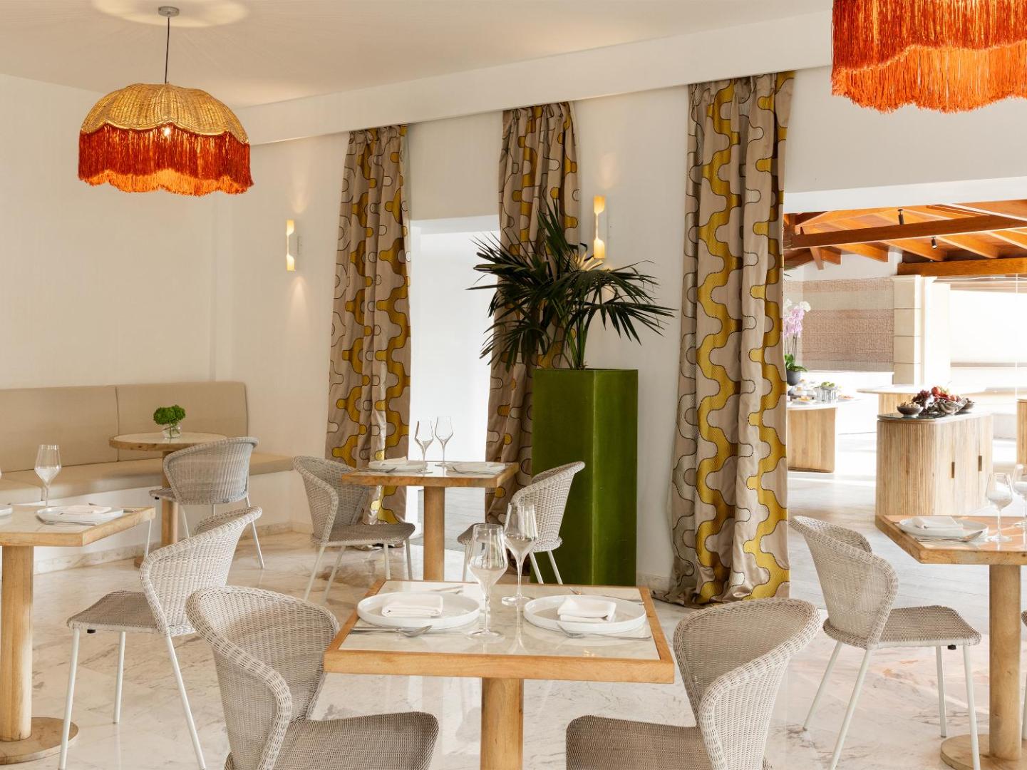 Grecotel Casa Adele restorāna interjers ar pītiem krēsliem, koka galdiem un stilīgiem oranžiem gaismekļiem gaišā, elegantā dizainā