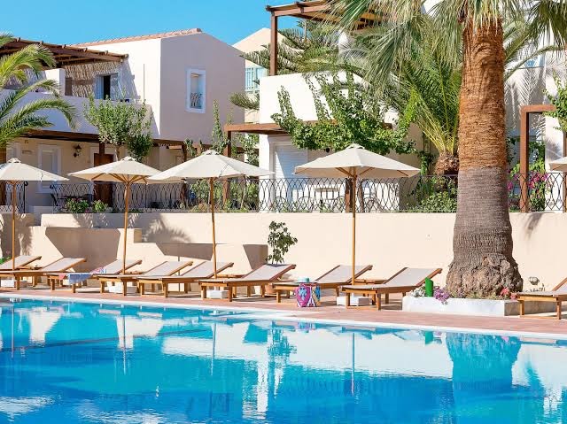 Tuvplāns ar sauļošanās krēsliem un saulessargiem pie Grecotel Casa Adele baseina Krētā — fonā baltas viesnīcas ēkas un palmas