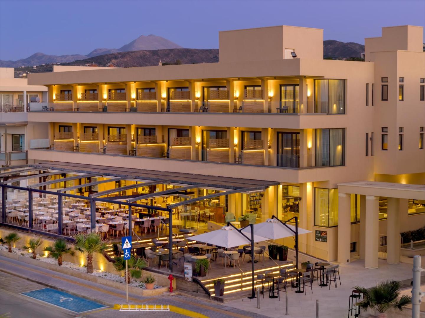 Atlantis Beach Hotel & Spa fasāde vakarā Retimno pilsētā Krētā — izgaismoti numuri, āra restorāns un kalnu panorāma fonā