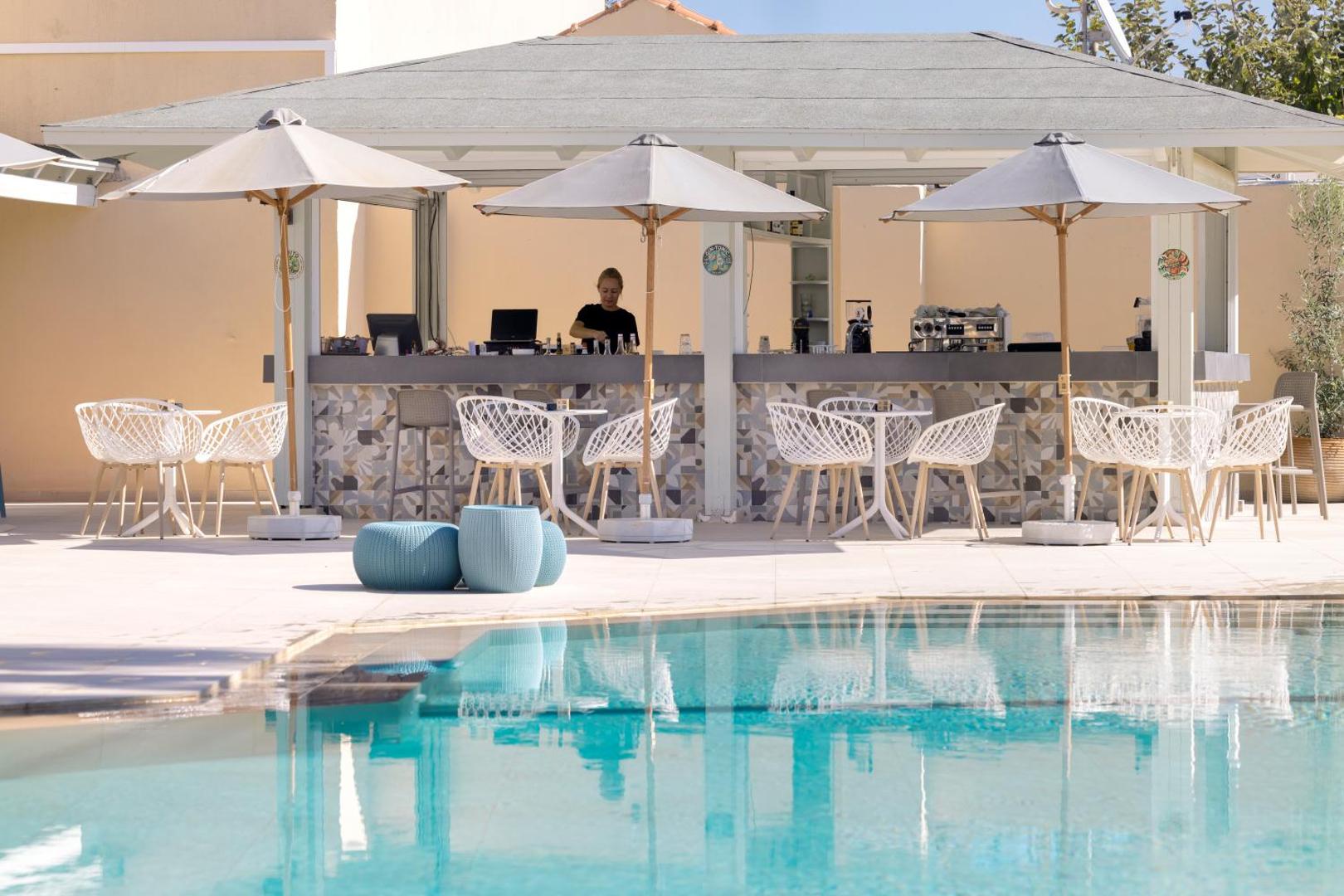 Atlantis Beach Hotel & Spa baseina bārs Retimno piekrastē ar moderniem krēsliem, saulessargiem un bārmeni pie letes saulainā dienā