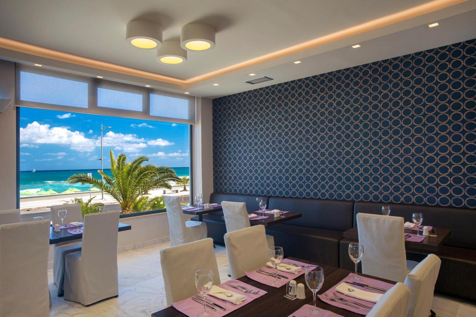 Atlantis Beach Hotel & Spa restorāna zāle ar skatu uz jūru — galdi ar violetiem akcentiem un panorāmas logu uz pludmali