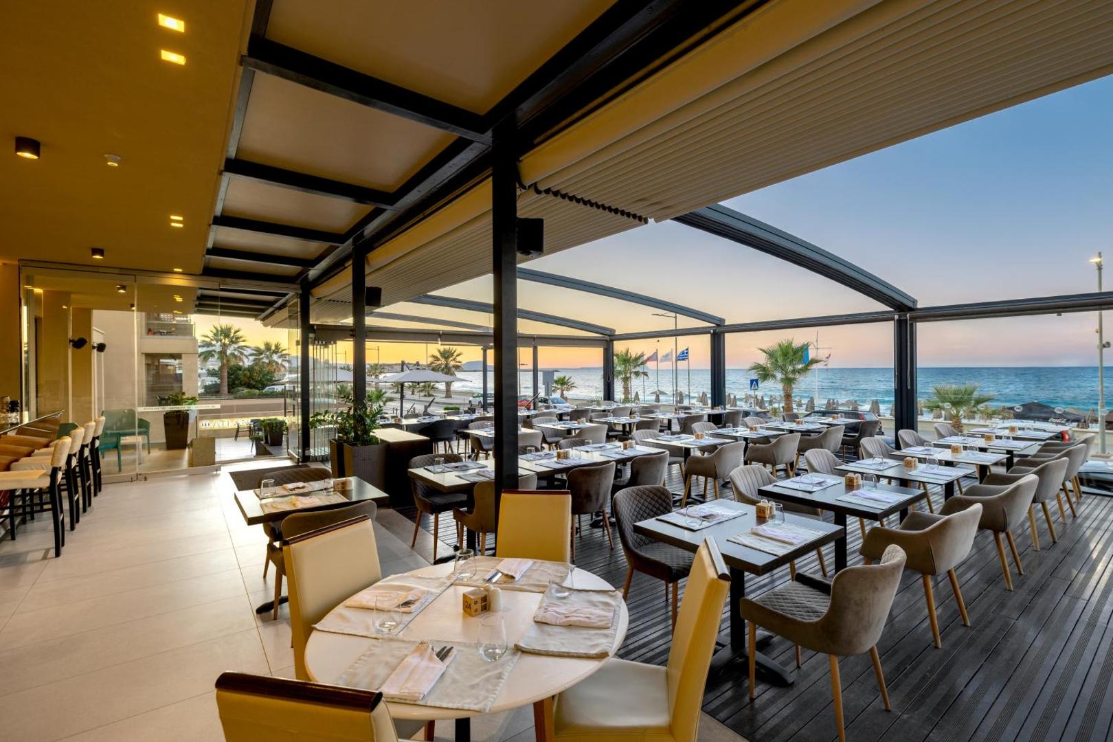 Atlantis Beach Hotel & Spa restorāna terase ar skatu uz jūru saulrietā, eleganti klātiem galdiem un modernu apgaismojumu