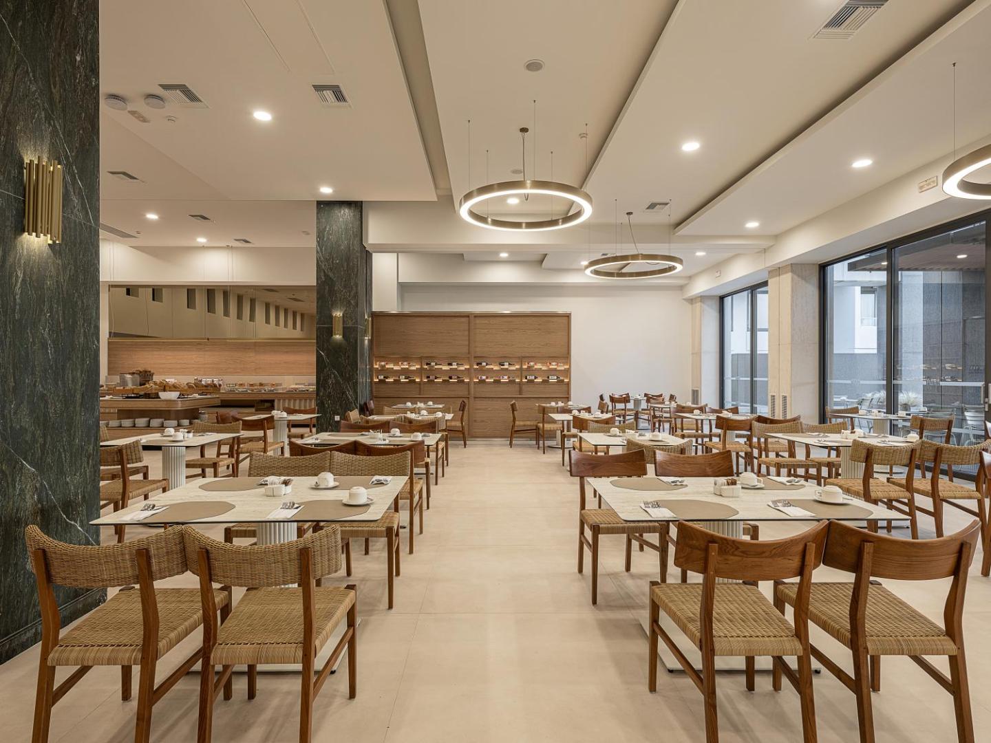 Mūsdienīga Avra Imperial Hotel brokastu zāle ar gaišu interjeru, koka mēbelēm un moderniem gredzenveida gaismekļiem pie griestiem