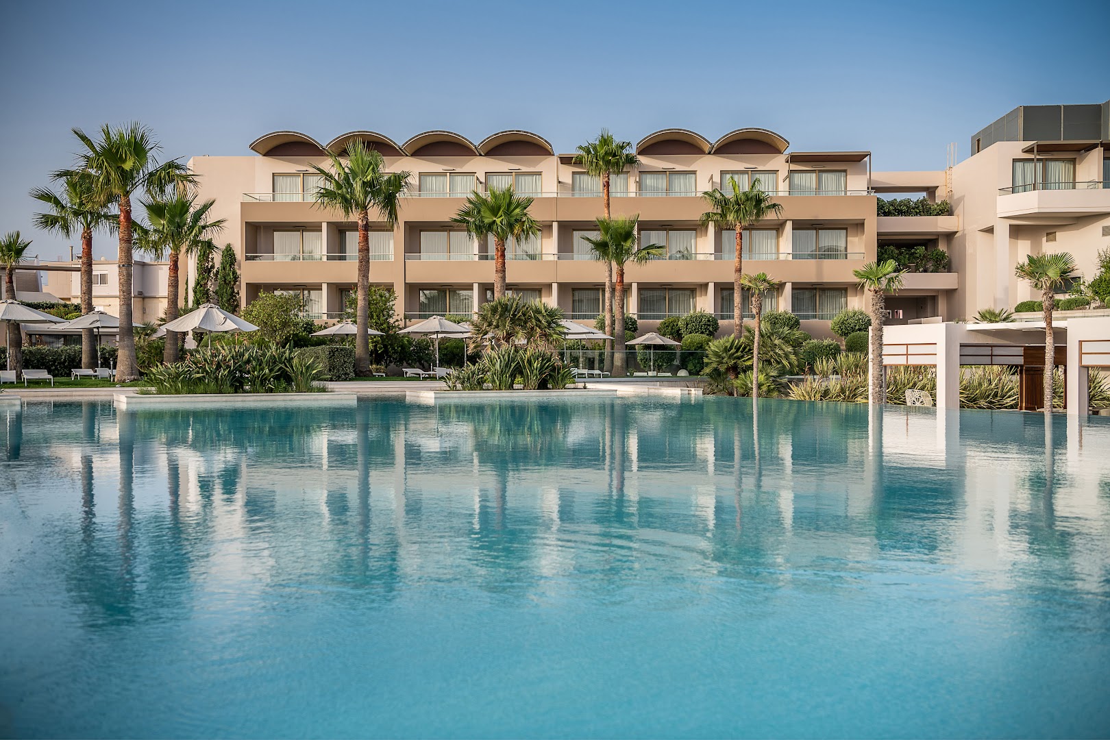 Avra Imperial Beach Resort galvenais baseins un viesnīcas fasāde Krētā — plaša atpūtas zona ar palmām un saulessargiem