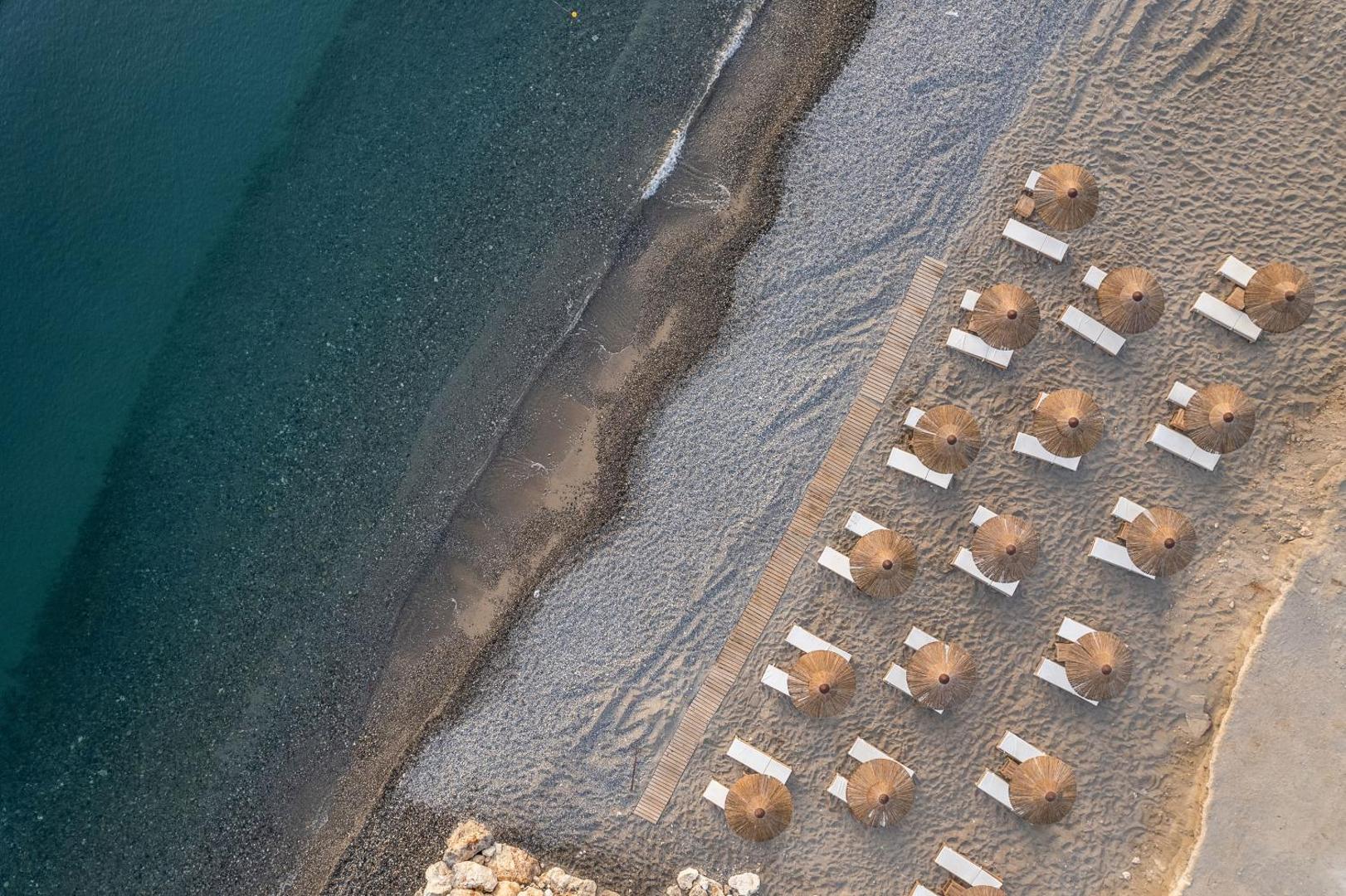 Aerofoto skats uz Avra Imperial Hotel pludmali — dzidrs jūras ūdens, oļu krasts un simetriski izvietoti salmu saulessargi ar zviļņiem
