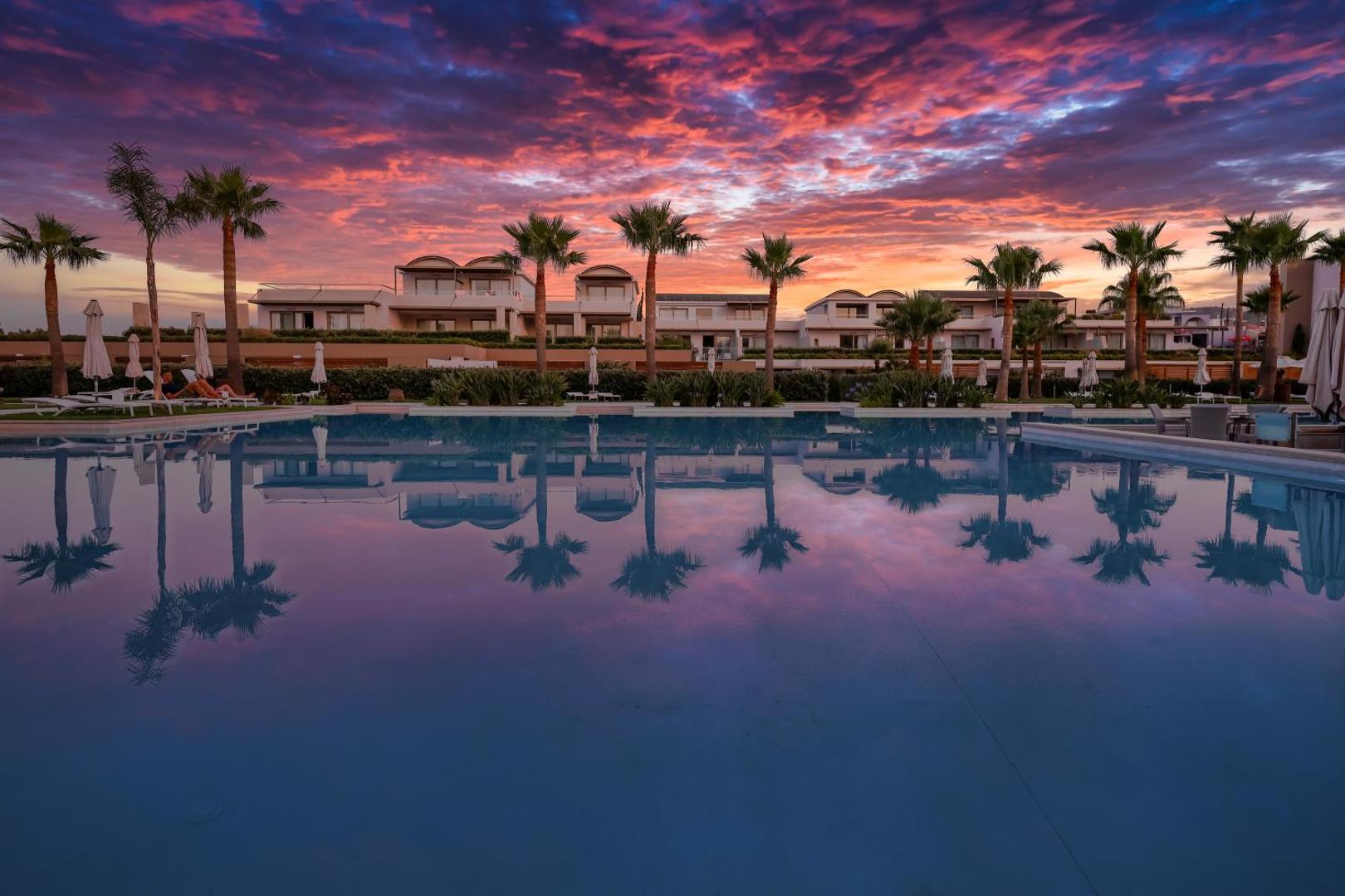 Avra Imperial Beach Resort baseins saulrieta laikā ar krāšņām debesīm un palmu atspulgiem ūdenī Krētas kūrortā