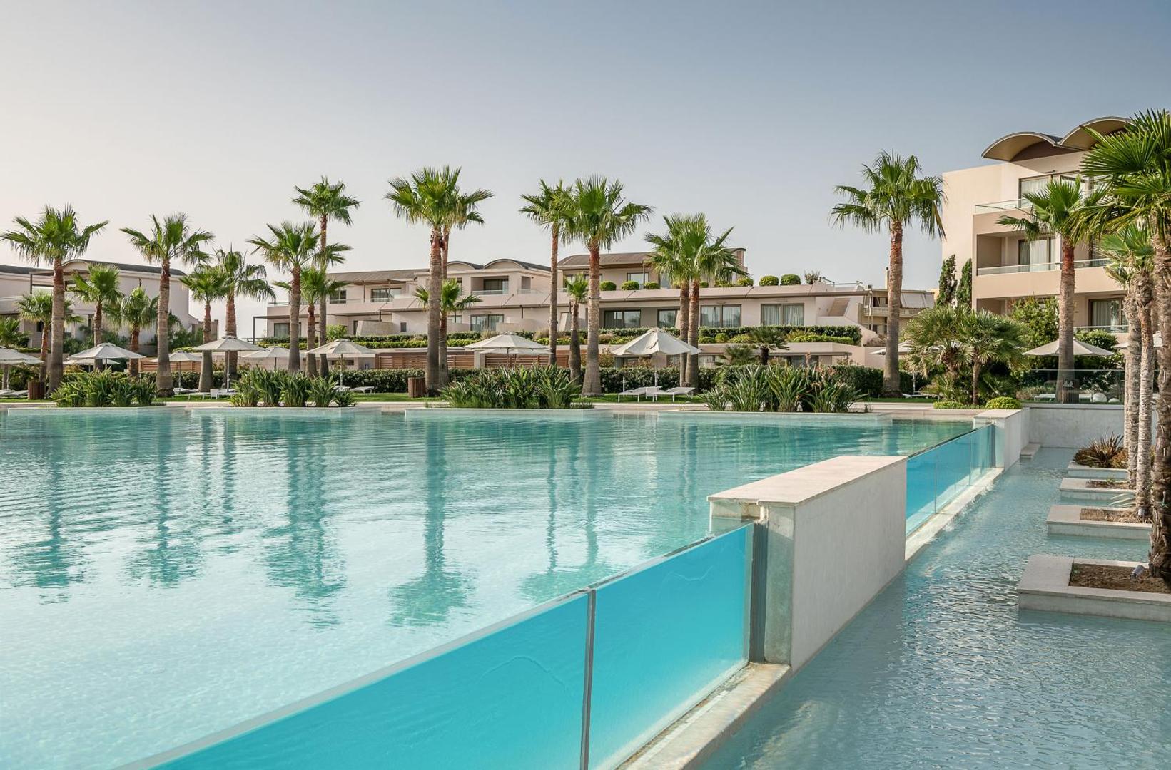 Avra Imperial Beach Resort modernais baseins ar stikla sienu un palmu rindu fonā Krētas salā
