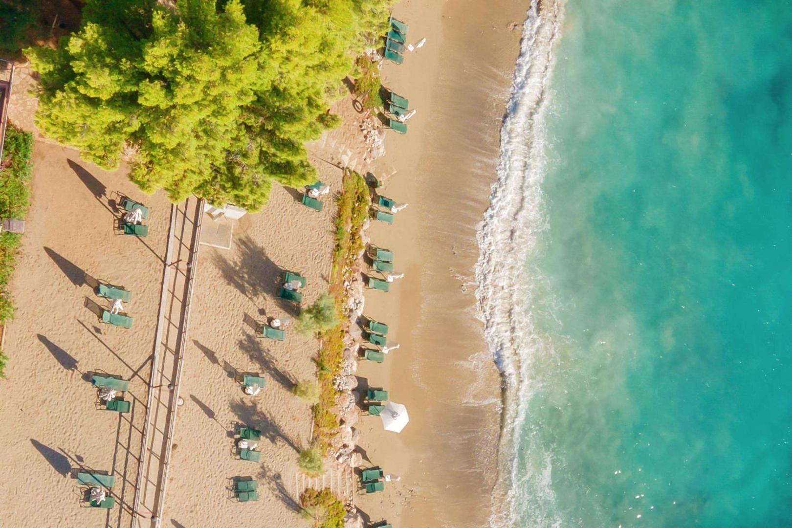 Candia Park Village privātā pludmale no putna lidojuma — smilšu krasts, zaļi koki, tirkīza ūdens un sauļošanās krēsli