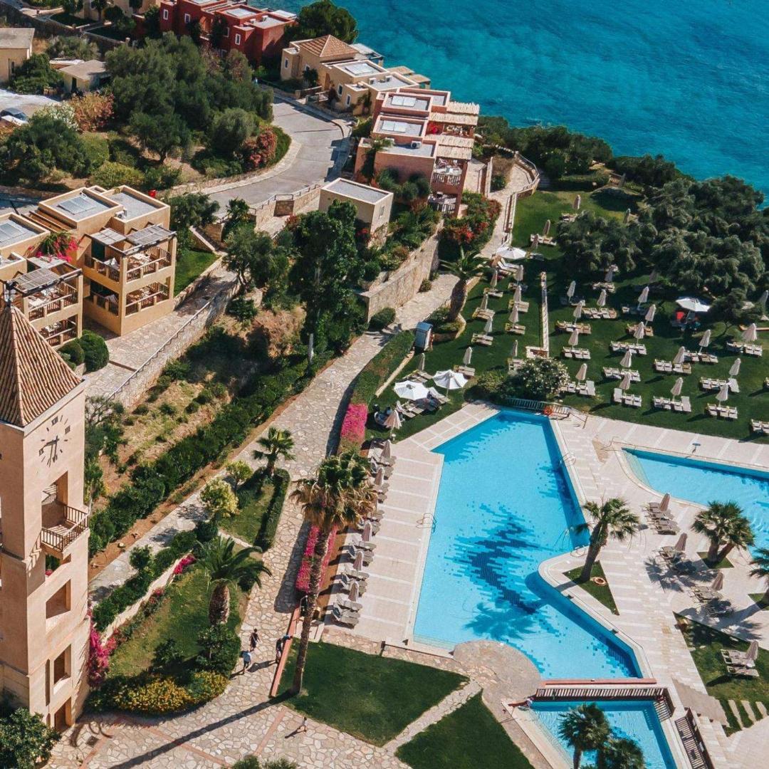 Candia Park Village teritorijas skats no augšas ar baseinu kompleksu, sauļošanās zonu zālienā un pulksteņa torni piekrastē.