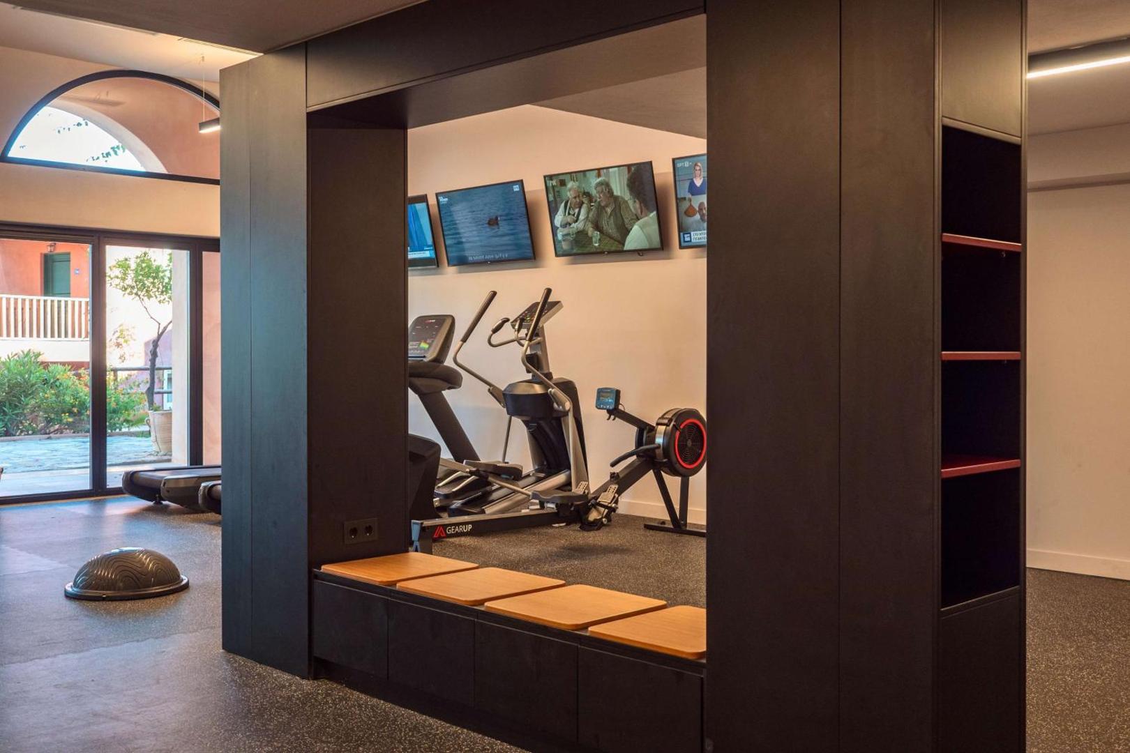 Candia Park Village moderns fitness centrs ar skrejceliņiem, airēšanas trenažieri un ekrāniem pie sienas gaišā numurā