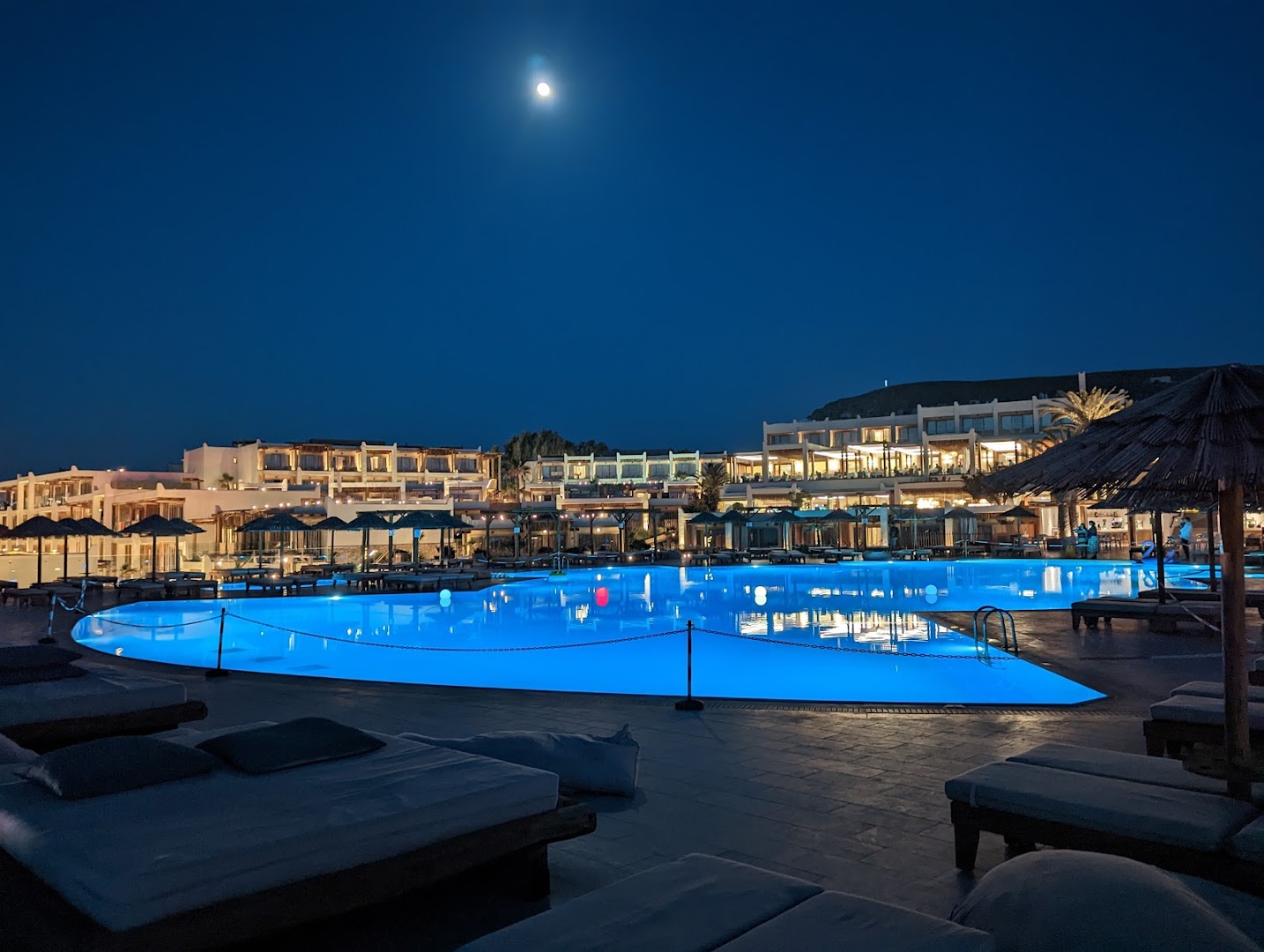Mitsis Rinela Beach Resort viesnīcas ēkas un izgaismots baseins nakts laikā zem pilnmēness — luksusa kūrorta atmosfēra Krētā