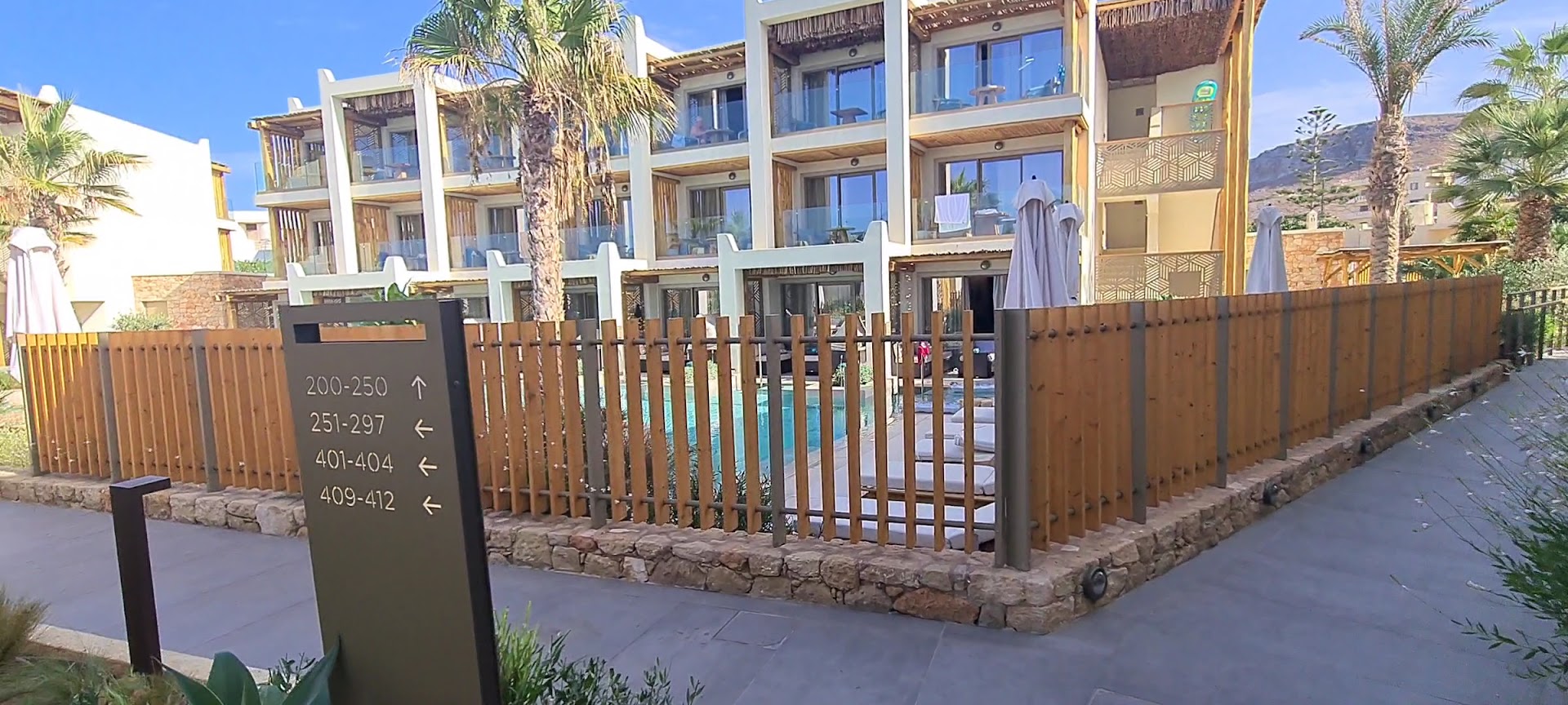 Mitsis Rinela Beach Resort norāžu zīme pie numuru korpusiem un baseina zonas ar palmām un koka elementiem