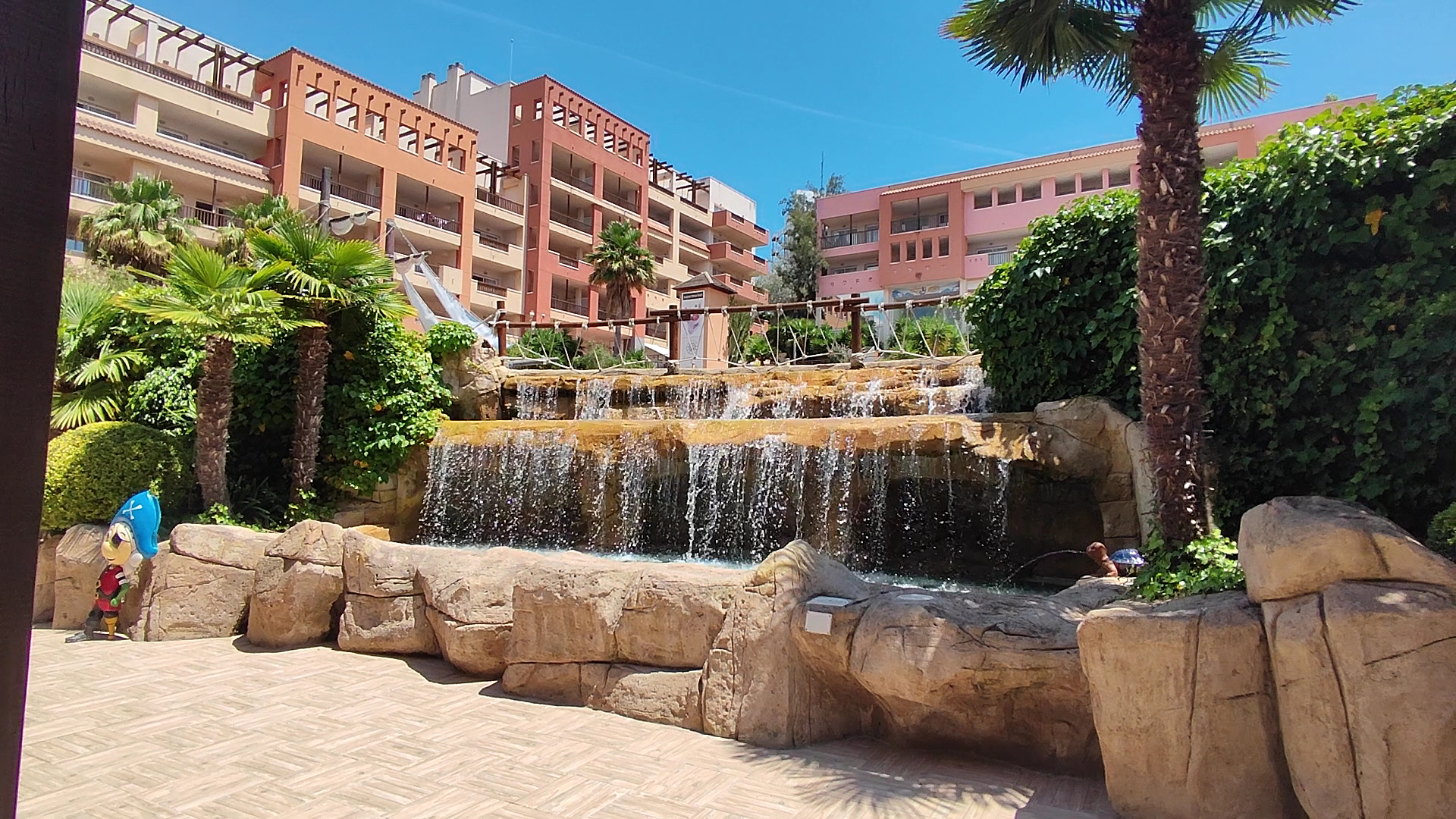 H10 Mediterranean Village dekoratīvais ūdenskritums un klinšu dārzs Salou reģionā — viesnīcas teritorijas labiekārtojums ar palmām un ēkām fonā