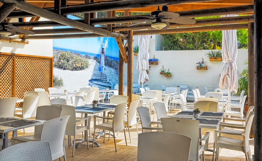 H10 Mediterranean Village viesnīcas āra terases restorāns ar baltiem krēsliem zem koka nojumes Kostadoradas piekrastē.