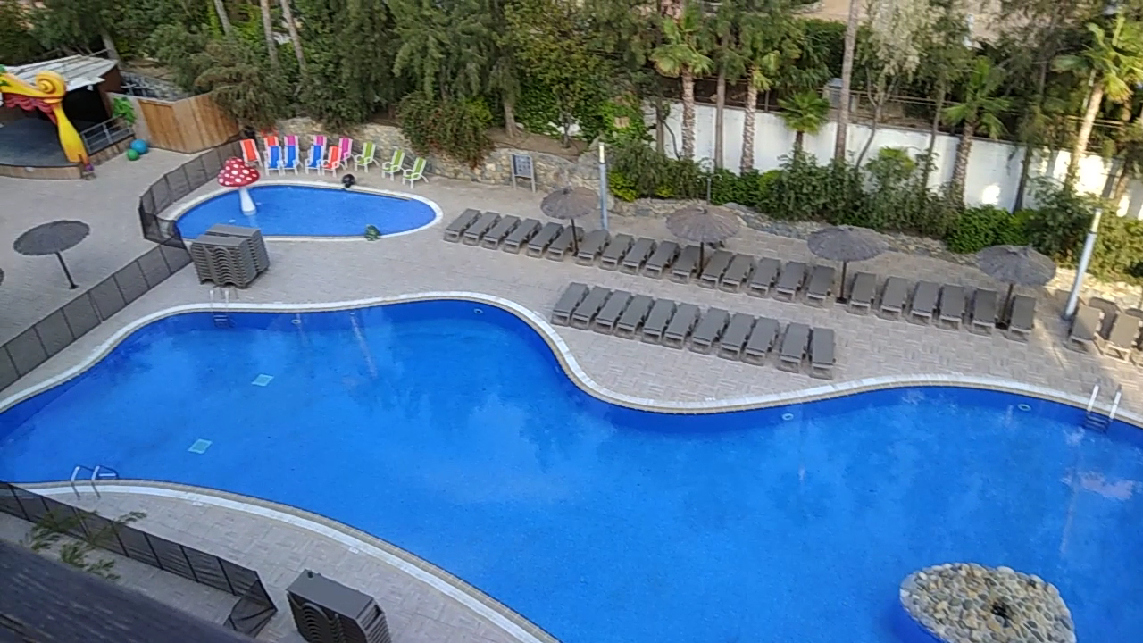 H10 Mediterranean Village āra baseinu zona Salovā ar bērnu baseinu, atpūtas krēsliem un saulessargiem priežu ieskautā teritorijā.